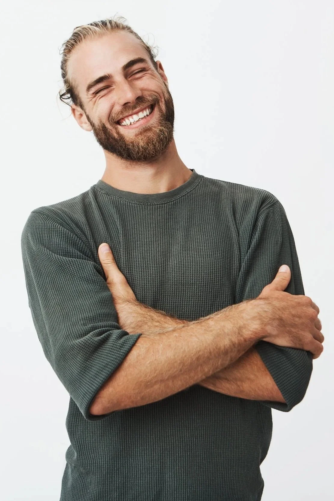 A smiling man with a beard and long hair tied back, crossing his arms, wearing a dark green long-sleeve shirt, against a plain white background.
