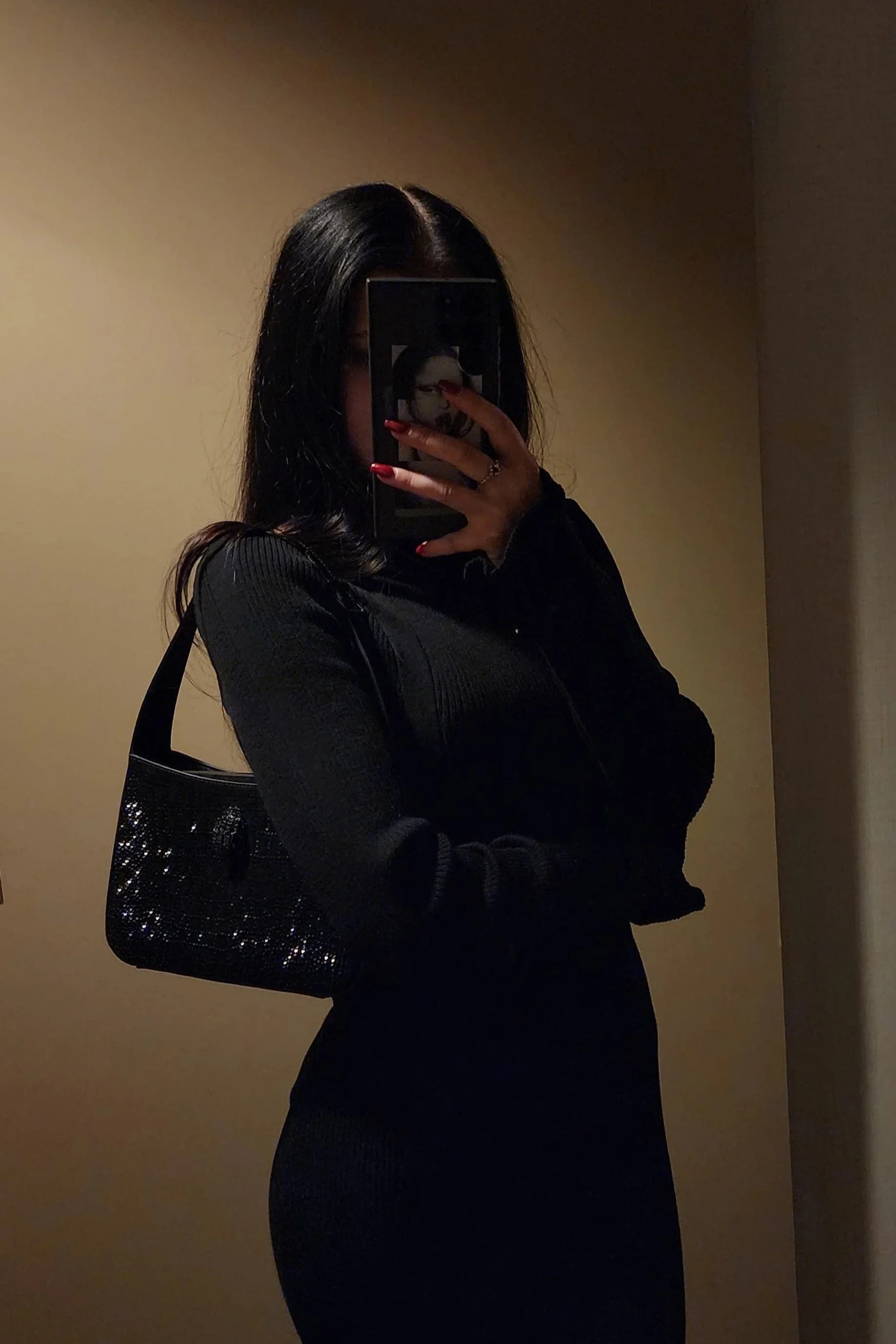 A woman with dark hair taking a mirror selfie in a dimly lit room, wearing a black long-sleeve top, a black skirt, and carrying a black sequined handbag.
