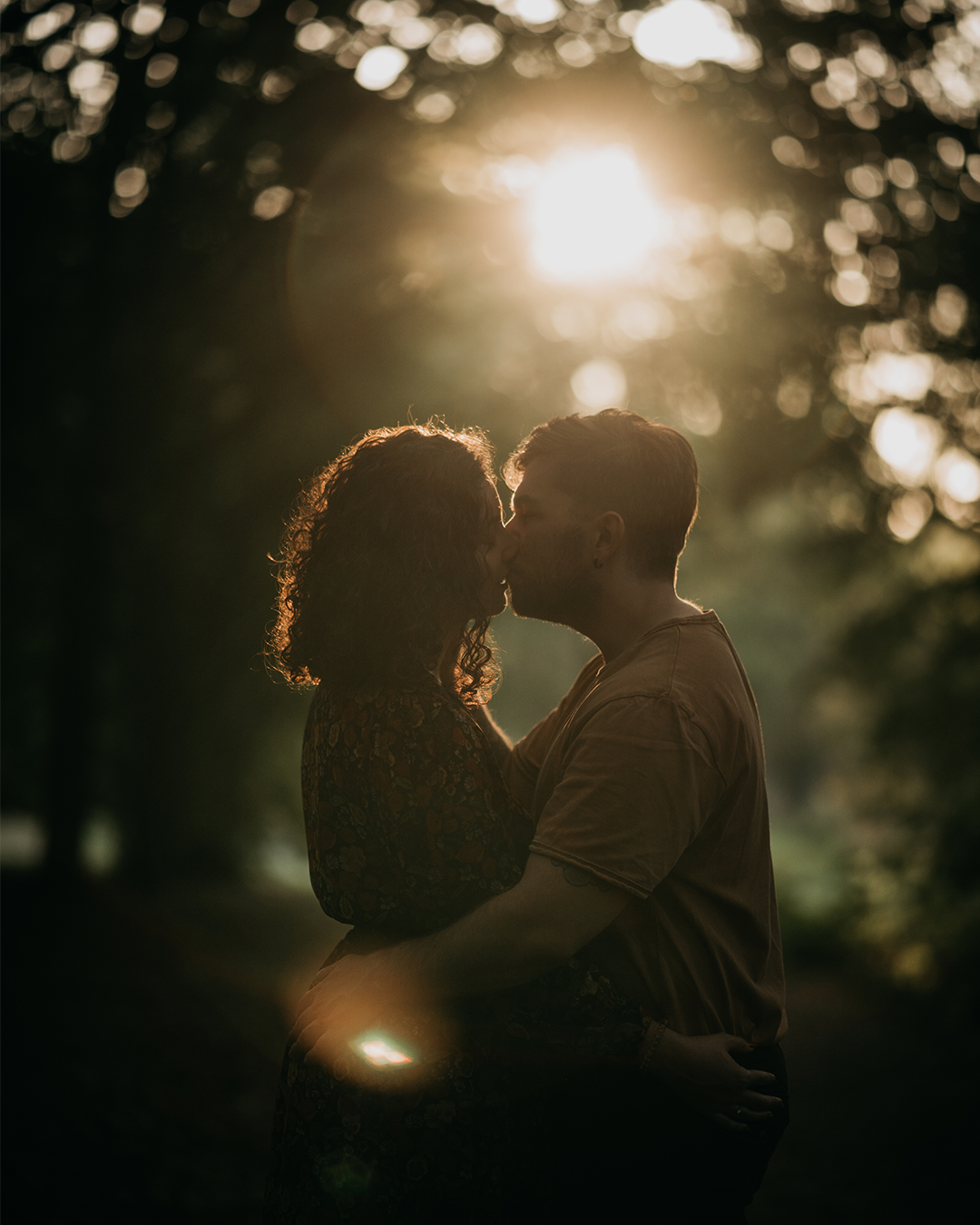 Un couple s'embrasse tendrement dans une forêt lors du coucher du soleil, entouré d'arbres avec la lumière dorée du soleil en arrière-plan.