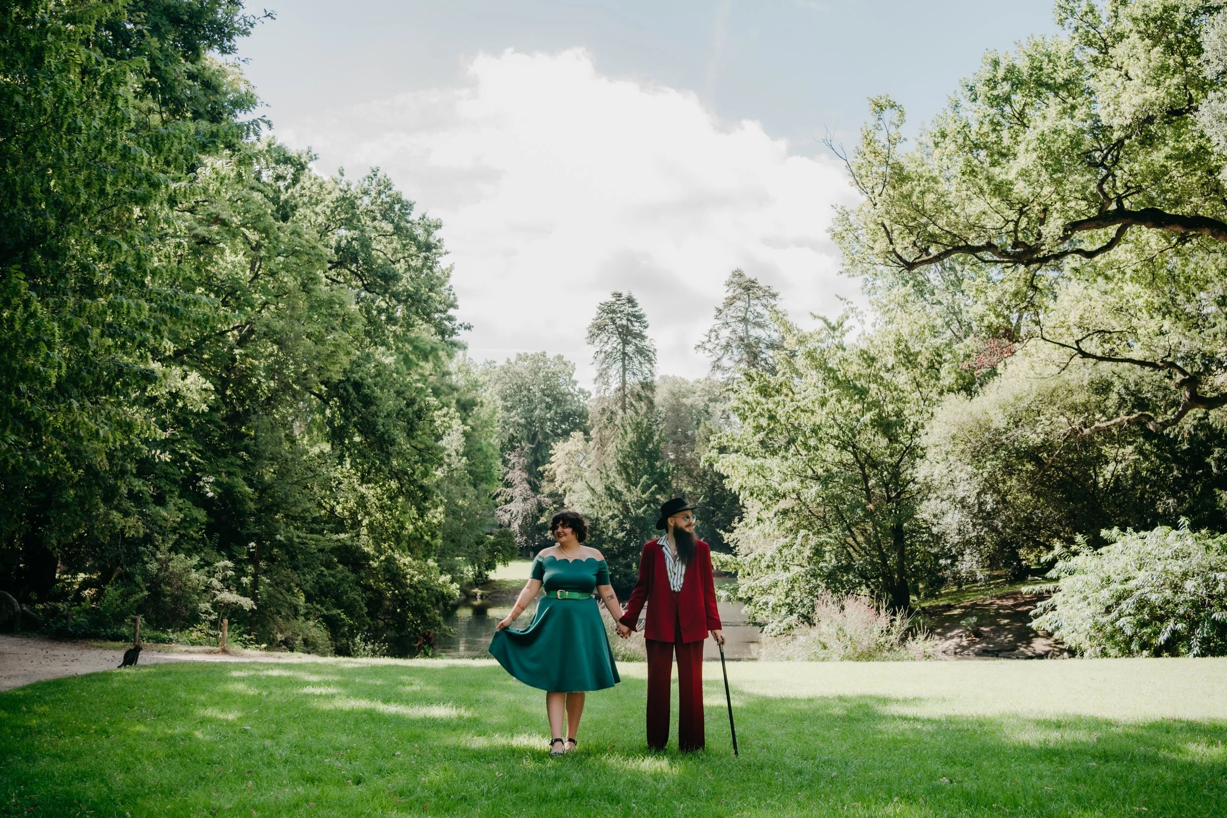 Un couple marchant dans un parc verdoyant avec des arbres et un ruisseau, portant des vêtements vintage colorés.