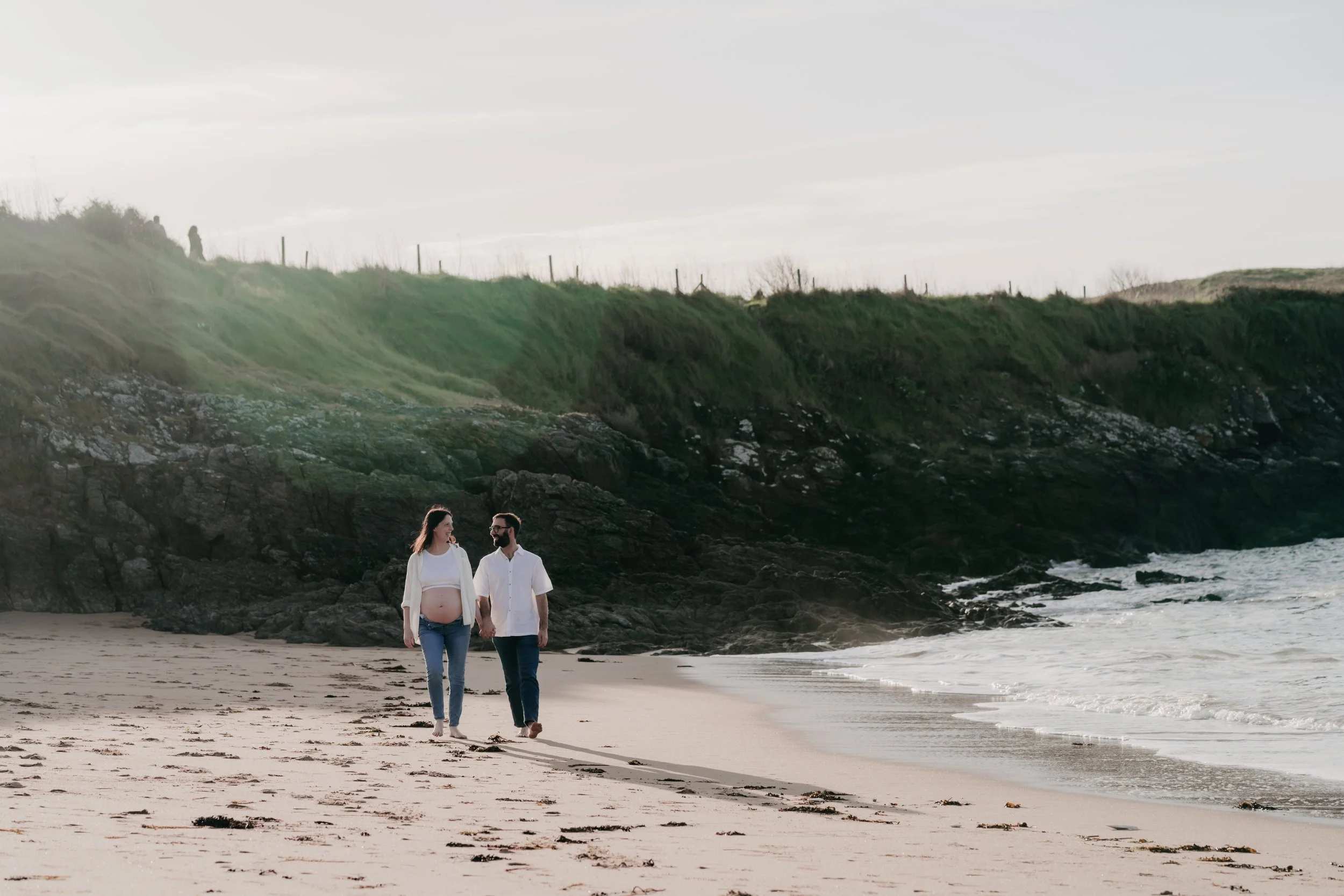 Un couple marche sur la plage près de l'océan, cheveux longs et vêtements décontractés, femme enceinte, arrière-plan de rochers et d'herbe verte sur une falaise, ciel nuageux.