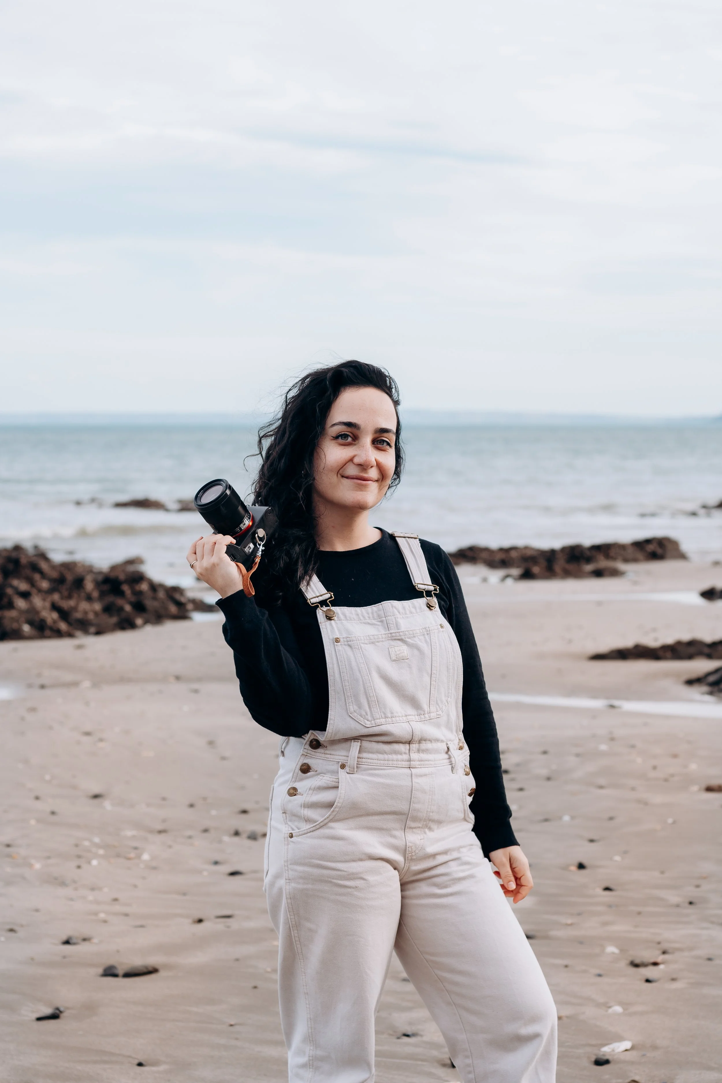 Une femme avec des cheveux noirs bouclés, portant un sweat noir et un pantalon beige, tient un appareil photo à la main sur la plage avec la mer en arrière-plan.