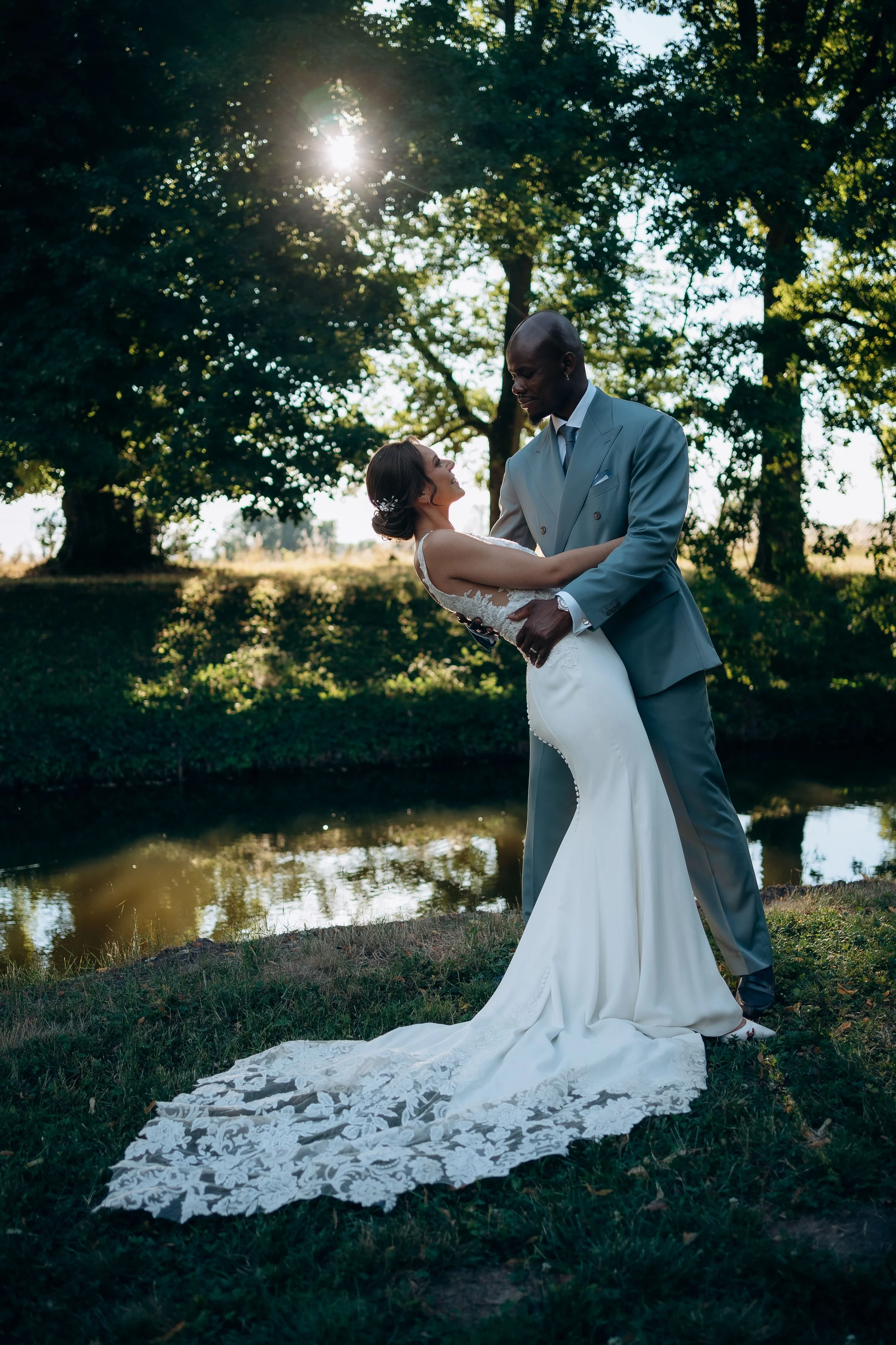 Un couple marié danse près d'une rivière dans un cadre naturel, avec des arbres en arrière-plan et le soleil derrière eux.