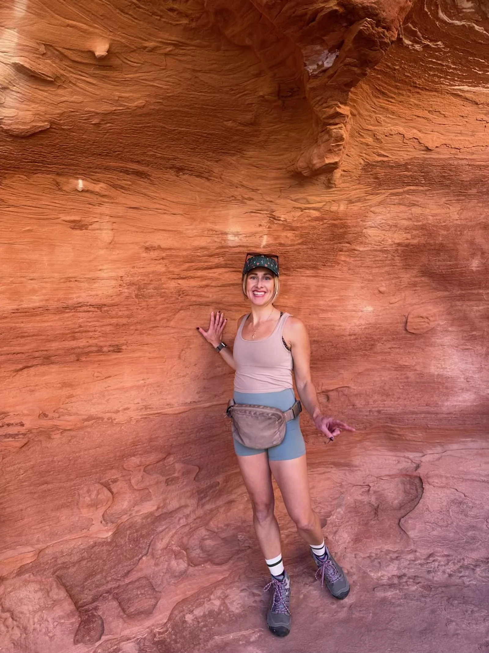 A woman smiling and standing inside a red sandstone canyon, wearing hiking gear, shorts, hiking boots, and a hat.