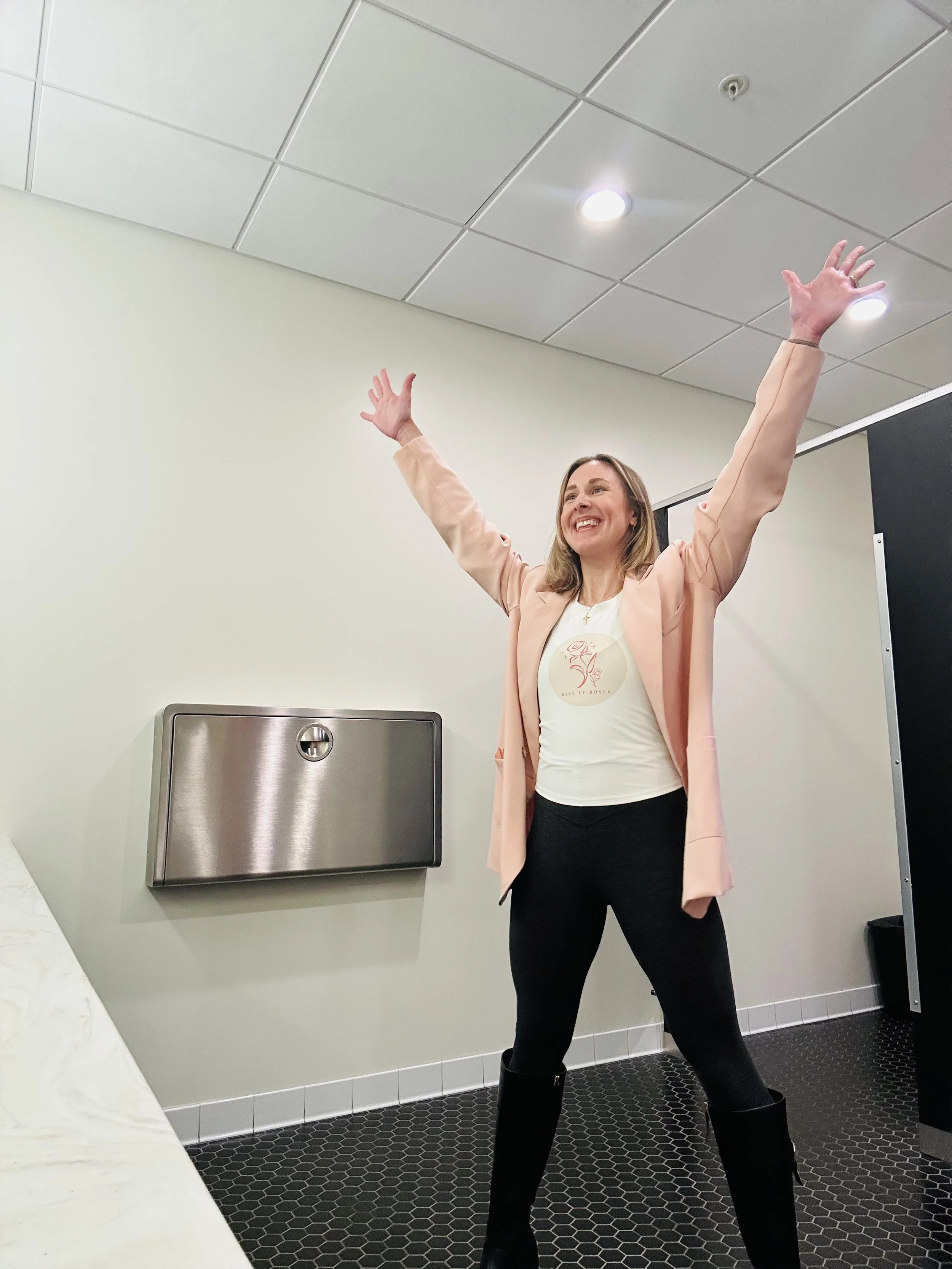 A woman with blonde hair, wearing a pink blazer, white t-shirt, black pants, and black boots, standing in a room with a gray wall, black tiled floor, her arms raised in celebration and smiling broadly.