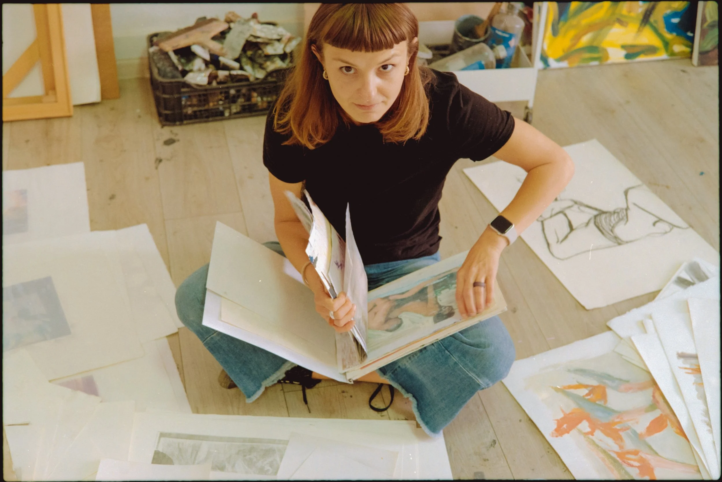A woman artist sitting on the floor with her legs crossed, looking up at the camera, surrounded by various artworks and sketches, holding a sketchbook.