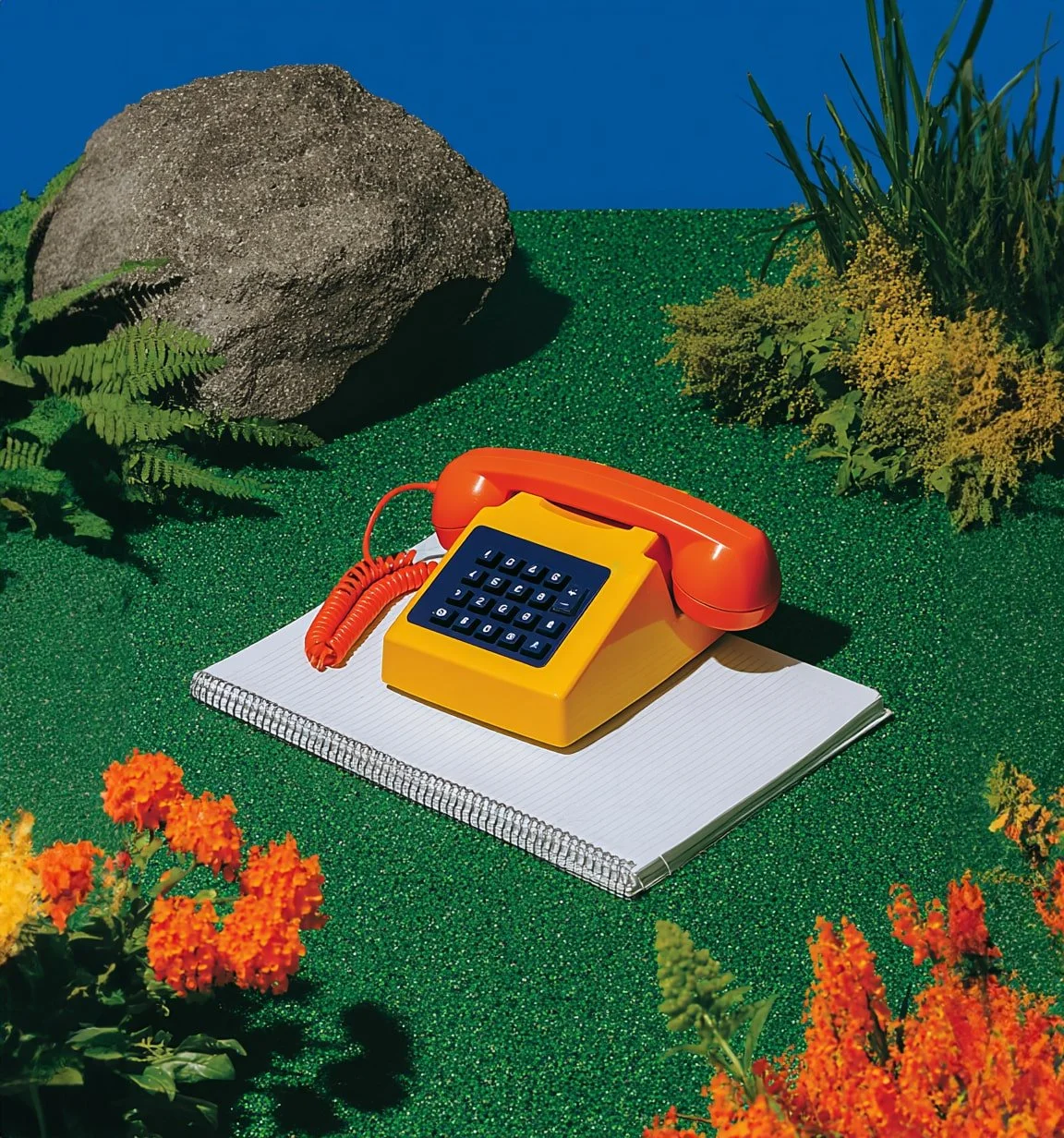 A vintage orange and yellow rotary phone resting on a spiral-bound notebook on green artificial grass with plants, rocks, and a blue sky background.