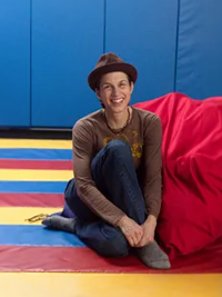 Person sitting on colorful floor with blue wall and red cushion, smiling and wearing a brown hat and casual clothes.