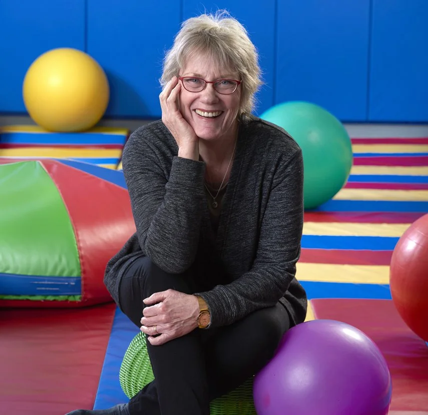 A smiling woman with short gray hair, red glasses, wearing a dark gray sweater, sitting on the floor of a colorful indoor gym with large exercise balls and a striped mat.