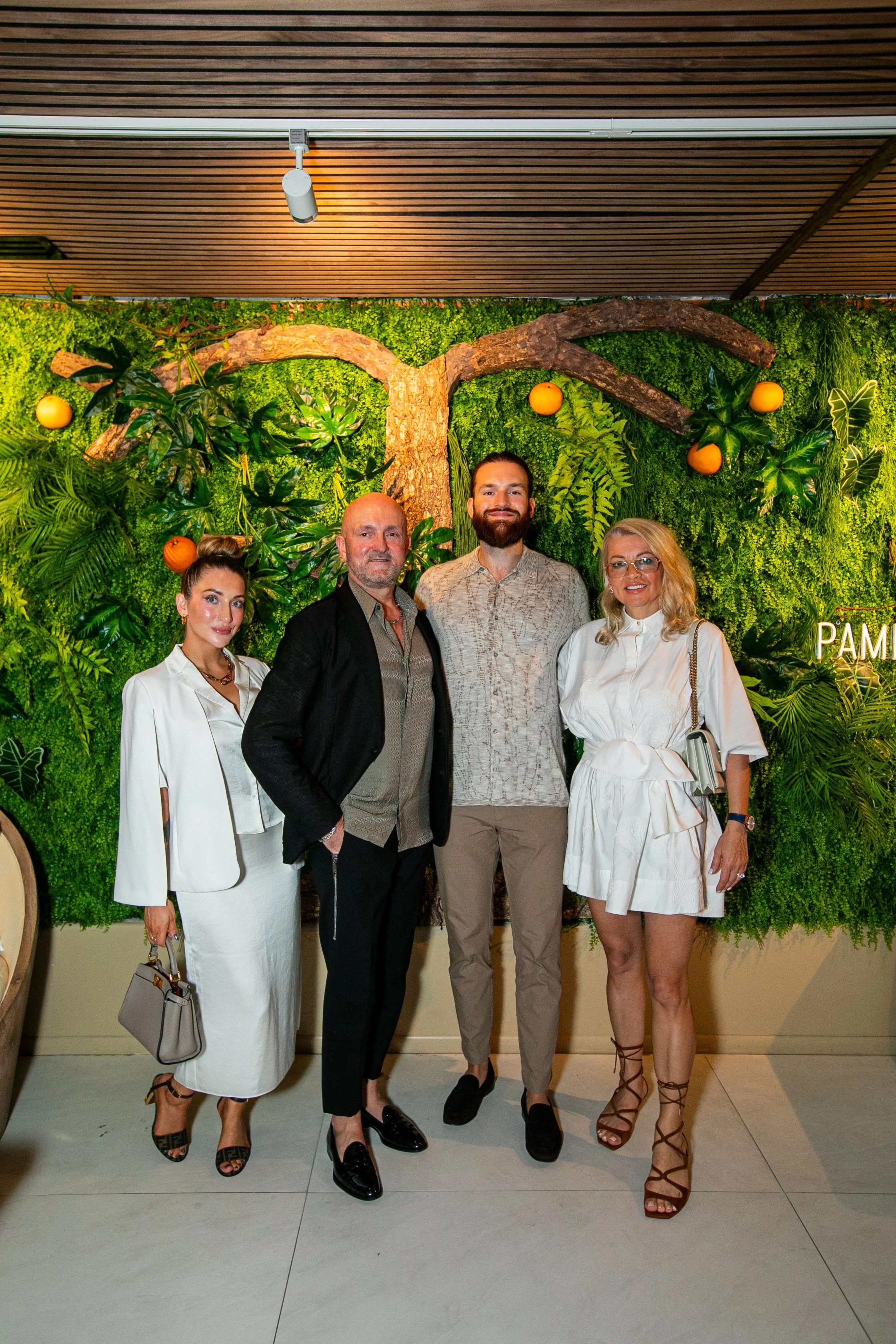 Four people stand in front of a green, leafy wall with a tree branch and orange fruit decorations, smiling at the camera.