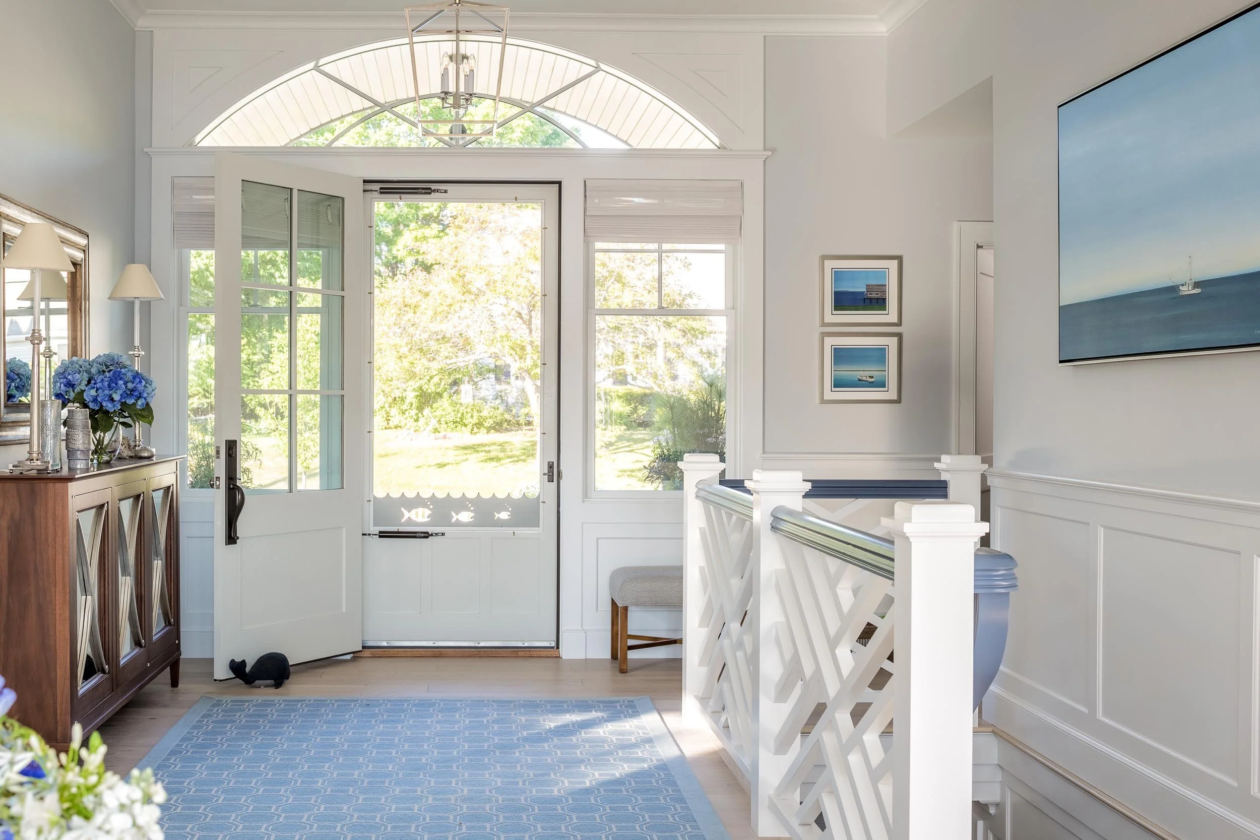 Bright entryway with white door and windows, wooden sideboard with decorative items, blue flowers, wall art, and a blue and white rug.