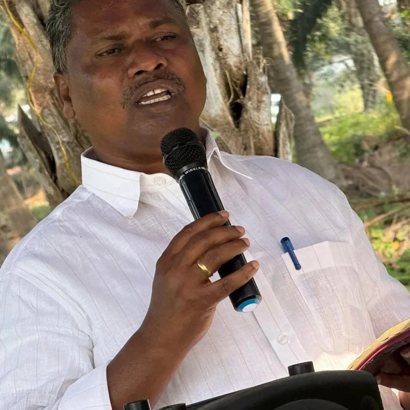 A man in a white shirt holding a microphone and a notepad, preaching with Bible in-hand outdoors.