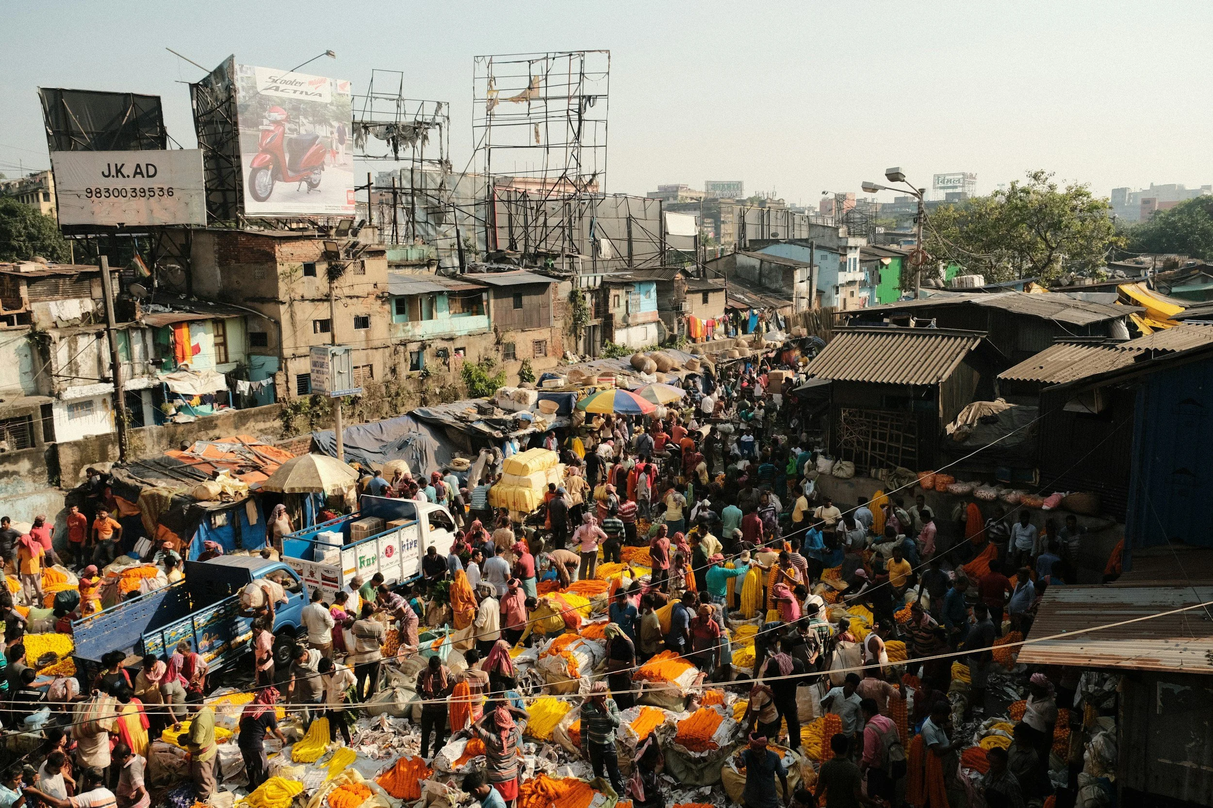 A busy outdoor marketplace in an urban area with many vendors and shoppers, surrounded by makeshift buildings and billboards.