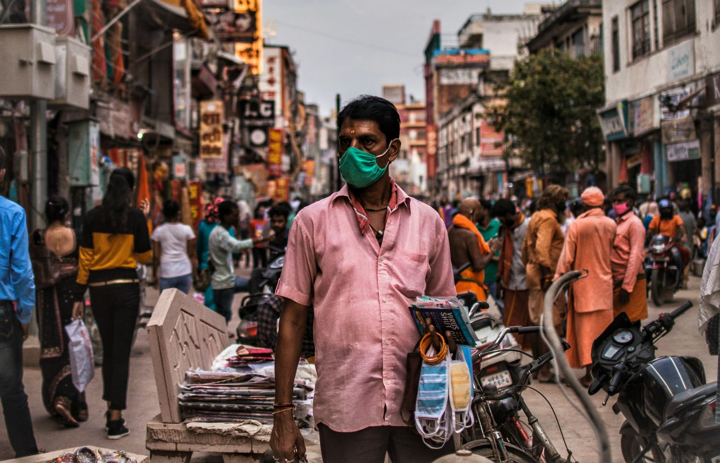 Man wearing a green face mask standing on busy street with shops and pedestrians, some wearing masks, in an urban market scene.