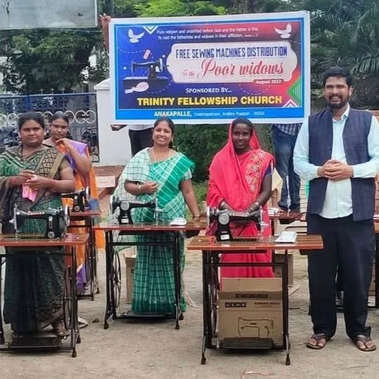 Group of women and a man standing behind sewing machines outdoors, with a sign above them advertising free sewing machines distribution.