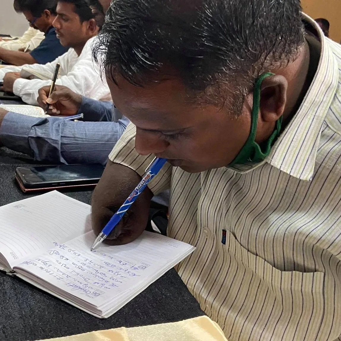 A man in a striped shirt writing in a notebook with a blue pen, sitting at a table during a meeting or class, other men in the background also taking notes.