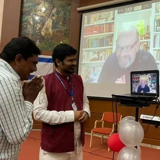 Two men, one with hands folded in a prayer gesture, face a screen displaying a man with glasses participating via video call. The scene appears to be a celebration or formal event with balloons and a decorated background.