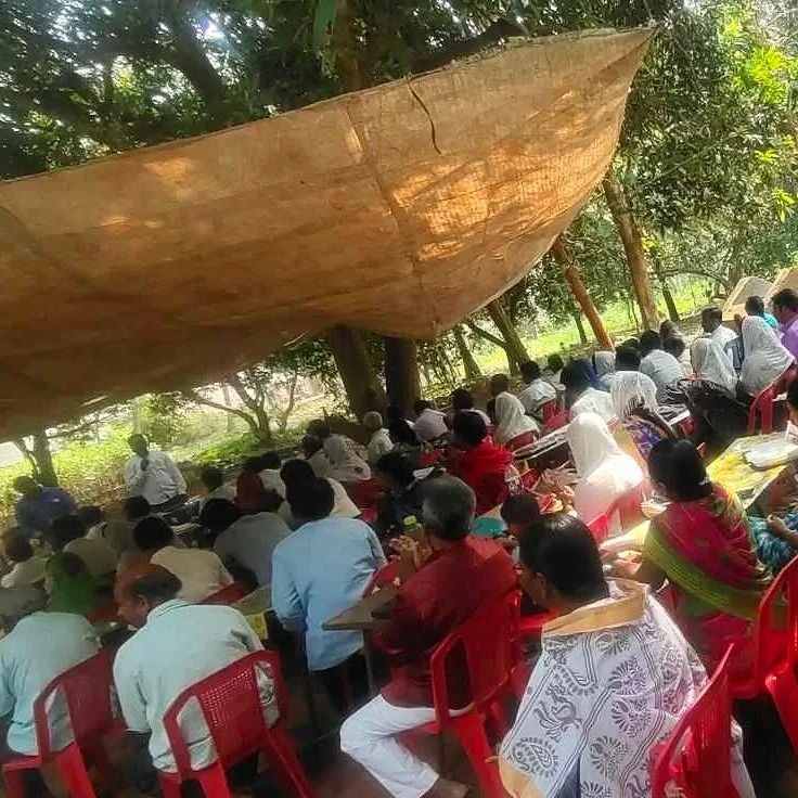 A large outdoor gathering of people seated under a makeshift tarp, sitting on red plastic chairs and tables in a shaded area with trees around.