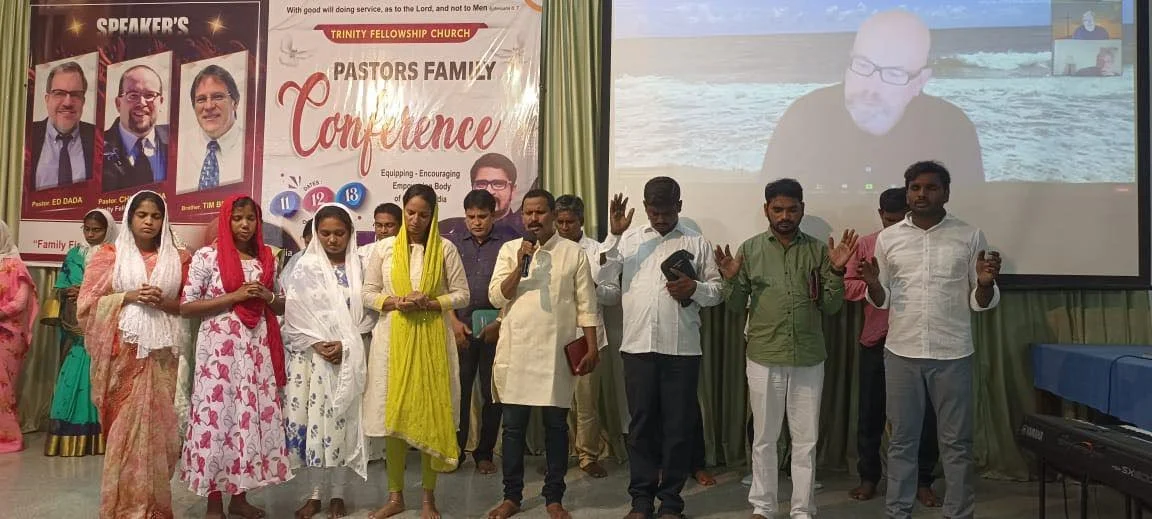 A group of people, including men and women, standing on a stage during a church conference with a banner in the background. Some have their eyes closed and are holding hands or with hands raised. There is a large screen showing a virtual speaker behind them.