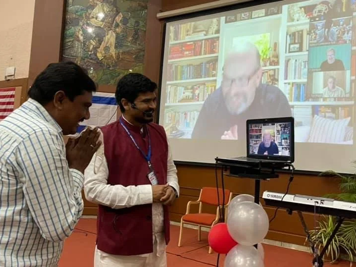 Two men are standing in front of a large projector screen displaying a video call with a man in a library. One man is bowing with hands pressed together; the other is smiling and clapping. There are decorative balloons and a keyboard in the room.