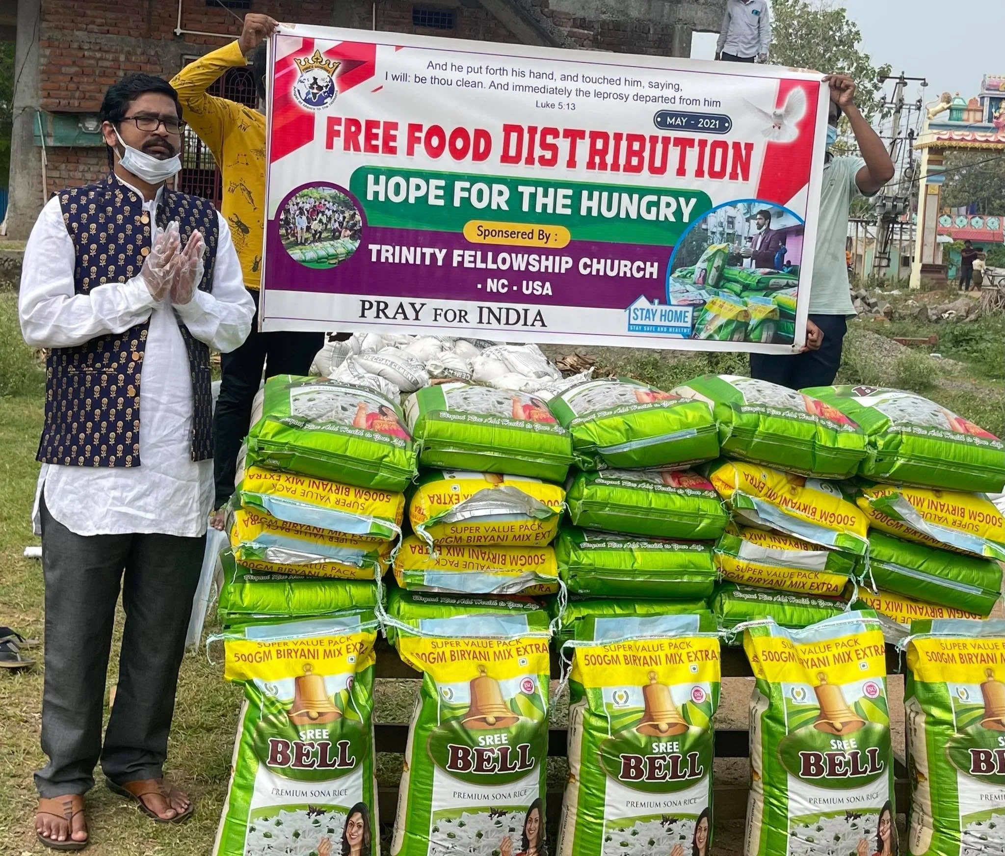 A man with glasses, wearing a mask, gloves, a white traditional outfit, and a blue patterned vest, stands with hands clasped in prayer next to a pile of rice bags. Behind him, two people hold a colorful banner promoting free food distribution, sponsored by Trinity Fellowship Church, with messages supporting India and encouraging staying home.