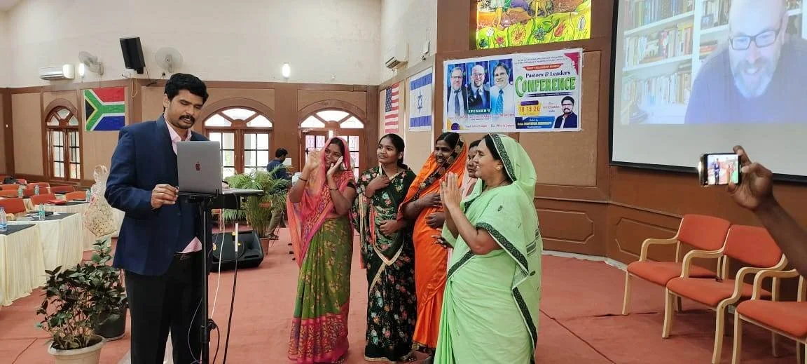 A group of women in traditional Indian sarees standing in a conference room with a man in a suit, participating in a virtual meeting projected on a large screen. A person is taking a photo with a smartphone.