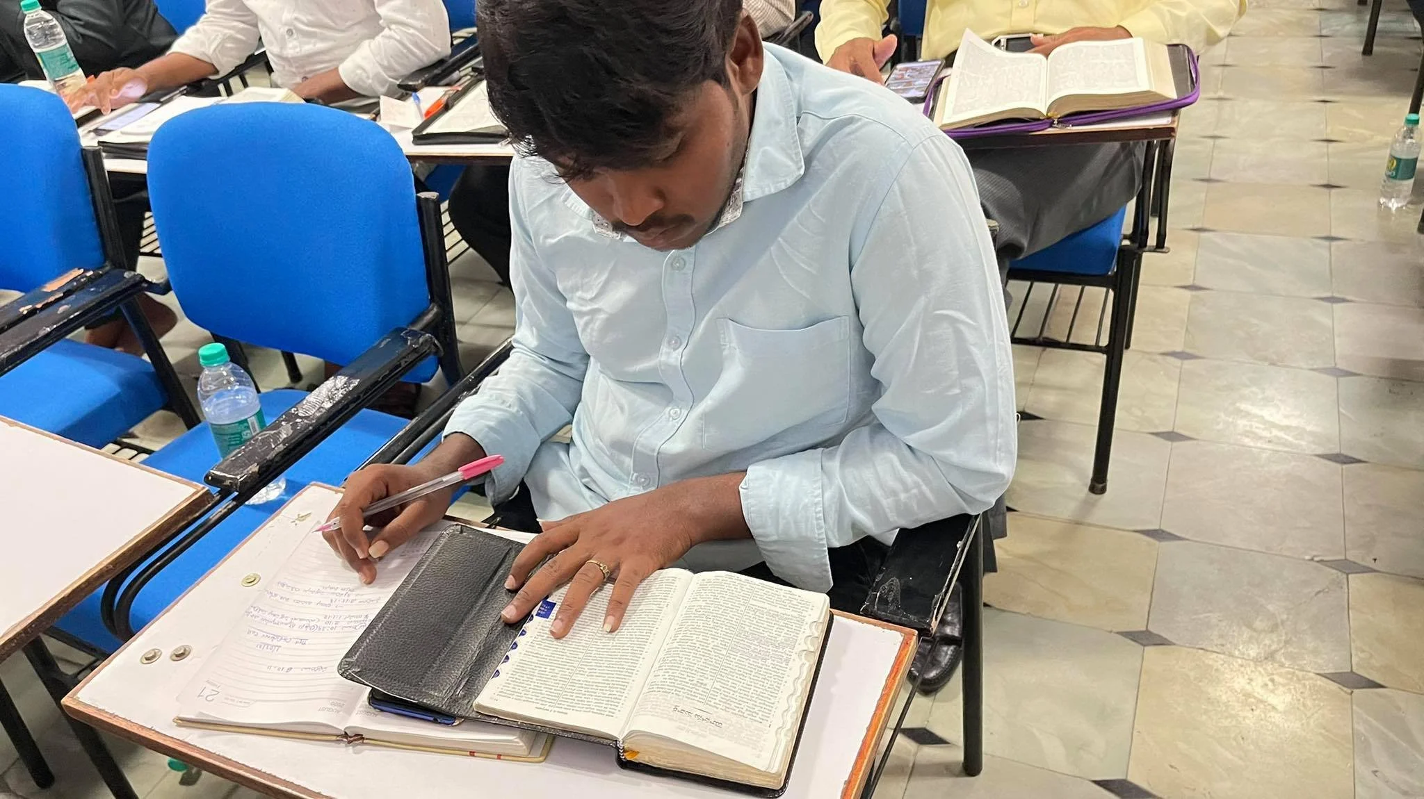 A man sitting at a desk in a classroom, reading a large open book and taking notes with a pink pen. There are other students and books in the background.
