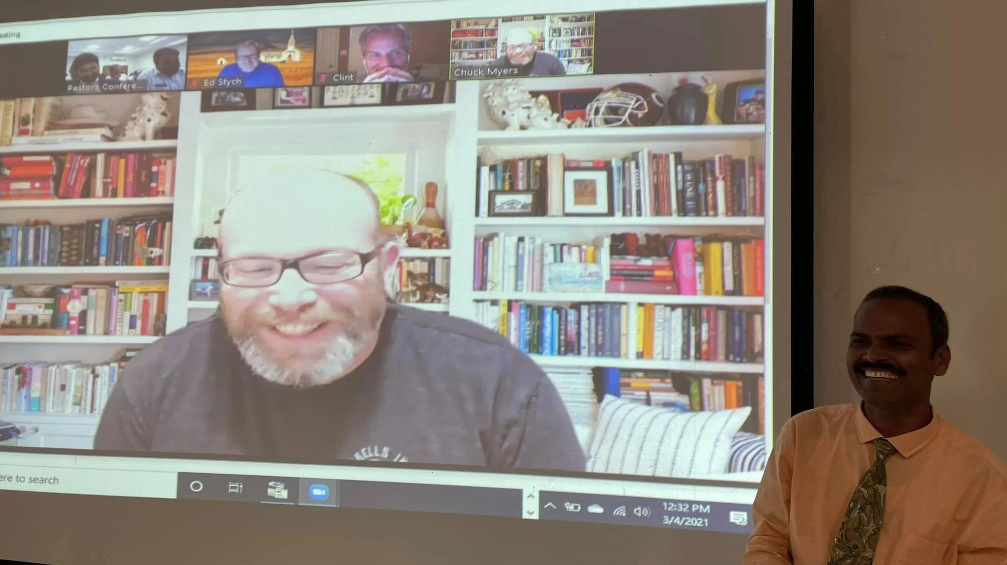 A virtual meeting displayed on a screen shows a smiling man with glasses in front of a bookshelf filled with books. An Indian man is seen leading the conference call, standing next to the screen and smiling.