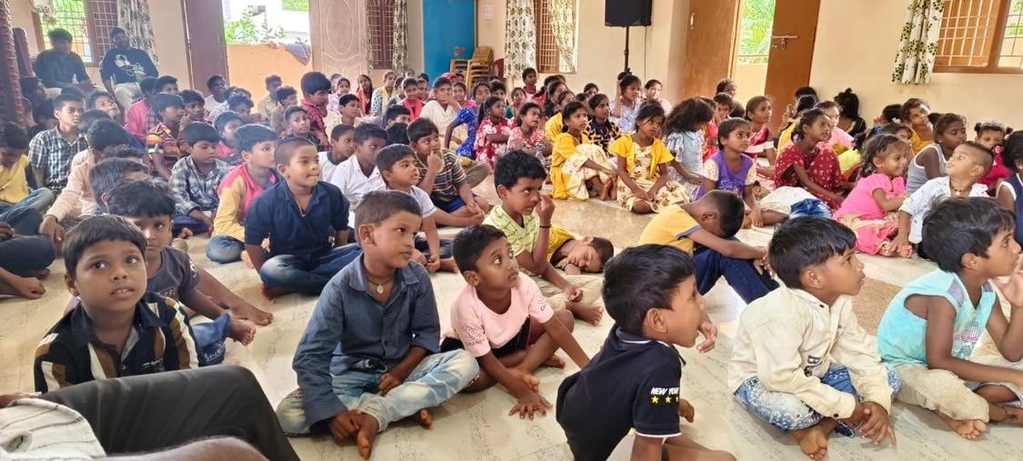 A large group of children sitting on the floor in a classroom or hall, paying attention to something in front of them.