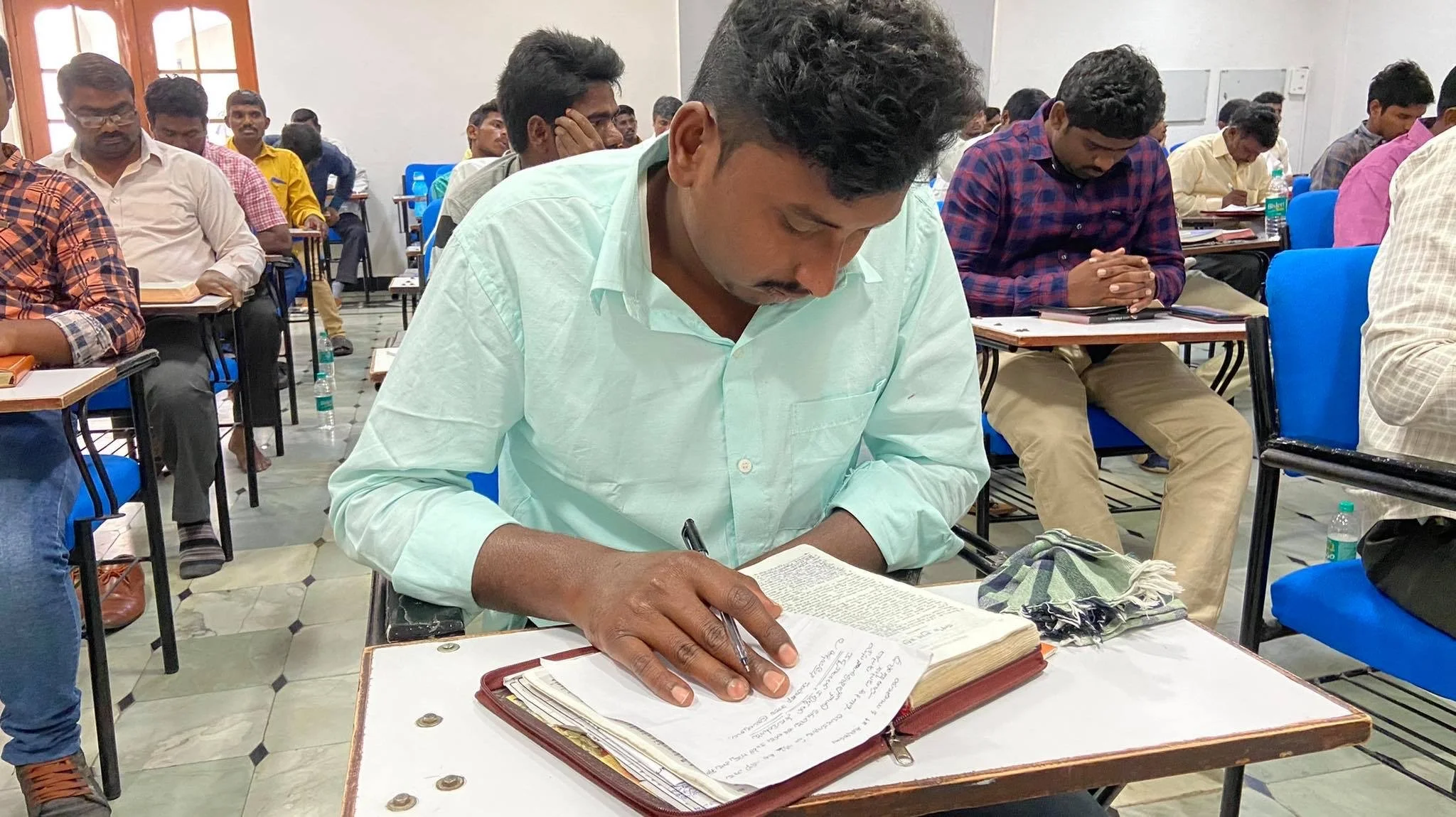 A man sitting at a desk reading and writing in a book during a classroom or seminar setting with other attendees in the background.