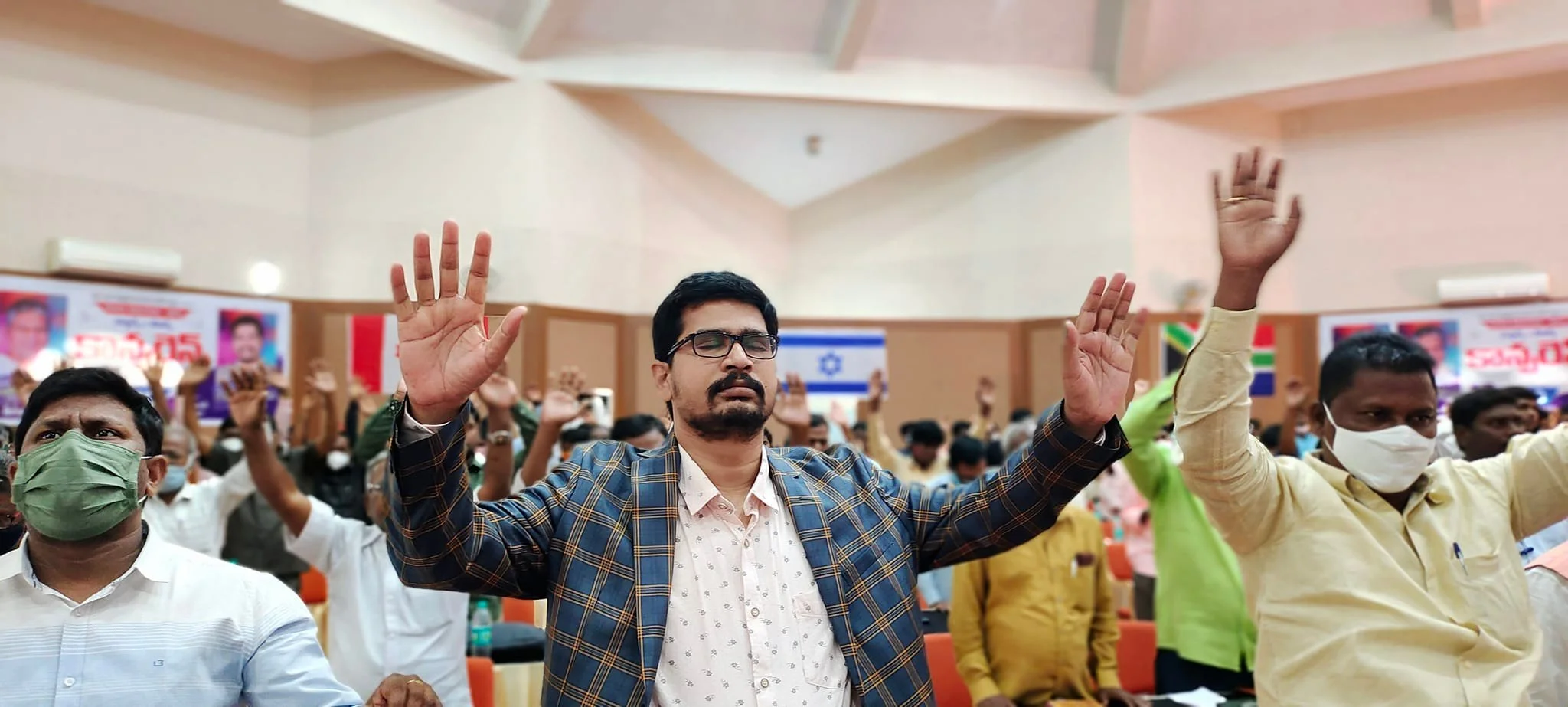 A group of people raising their hands in worship. The central figure is a man with glasses wearing a checkered blazer and a white shirt, with his eyes closed in prayer.