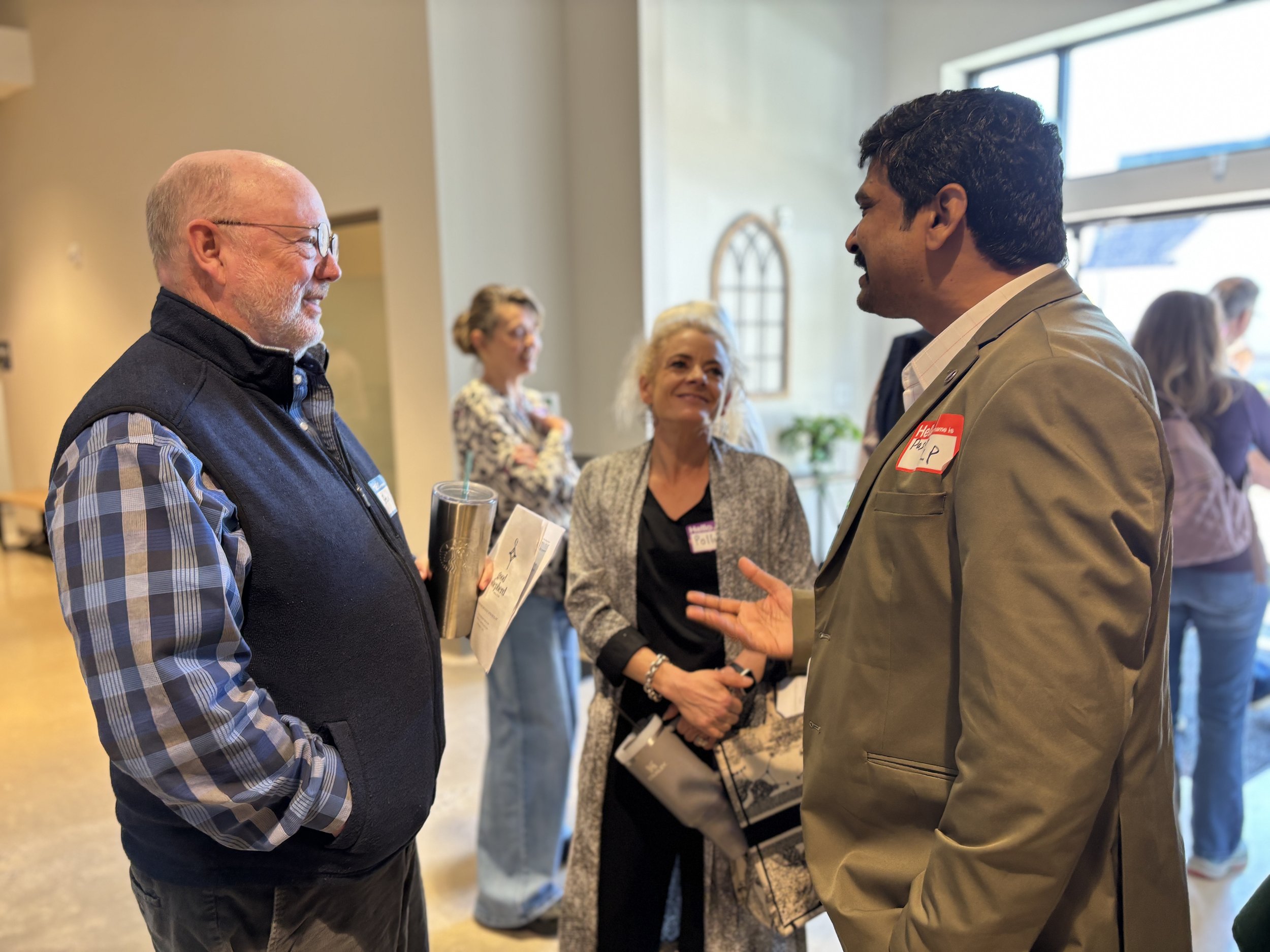 Two men and two women engaged in conversation at a church partner event in the USA. The man on the left has a beard and glasses, wearing a plaid shirt and a black vest. The man on the right has dark hair, wearing a beige blazer with a red name tag.