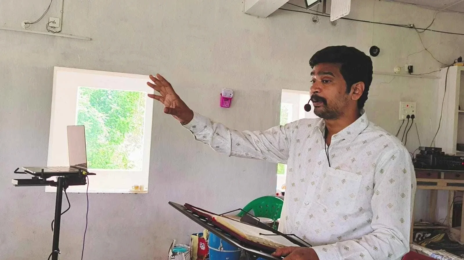 Man wearing a white shirt with a subtle pattern, speaking with a headset microphone, gesturing with his left hand in a room with white walls, a small window showing green trees outside, a laptop on a stand, and various electronic equipment in the background.