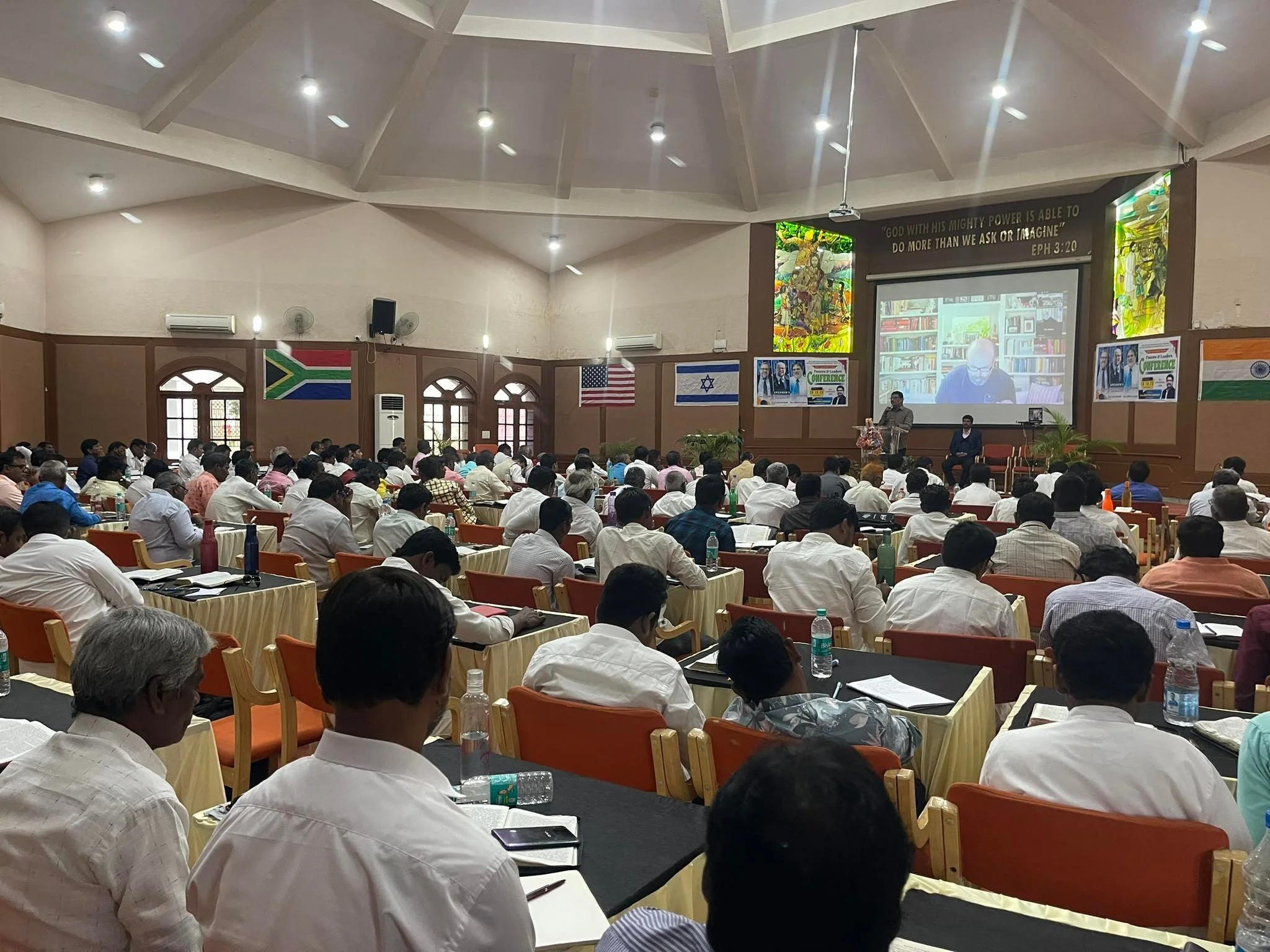 A large conference room filled with mostly men seated at tables, listening to a speaker at the front.  A large screen shows a virtual speaker in a library background.