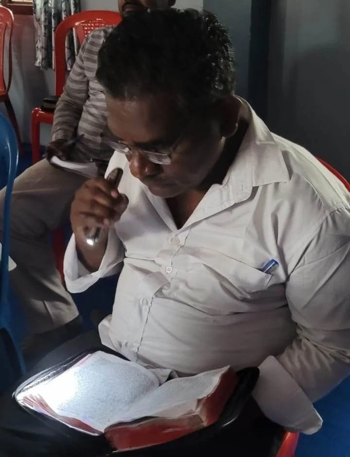A man wearing glasses and a white shirt sits in a dark room reading a Bible with the help of a flashlight.