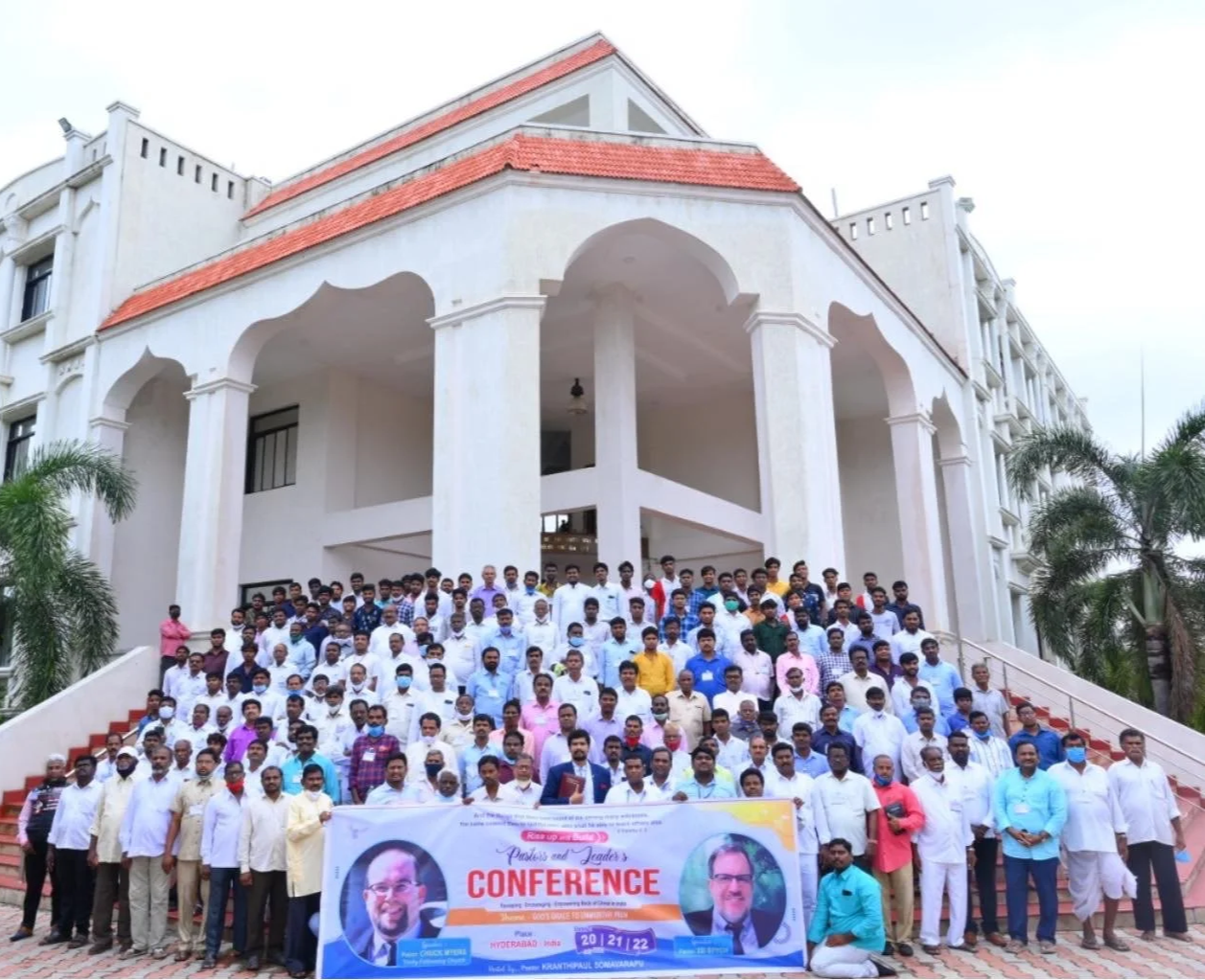 A very large group of men gathered in front of a white building, holding a conference banner with photos of teachers and co-founders, Ed Stych and Chuck Myers.