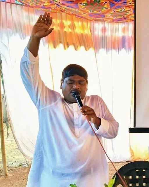A man in white traditional attire holding a microphone with his eyes closed, raising his right hand, and appearing to be praying or singing passionately. Light-colored curtains and a colorful cloth ceiling are in the background.