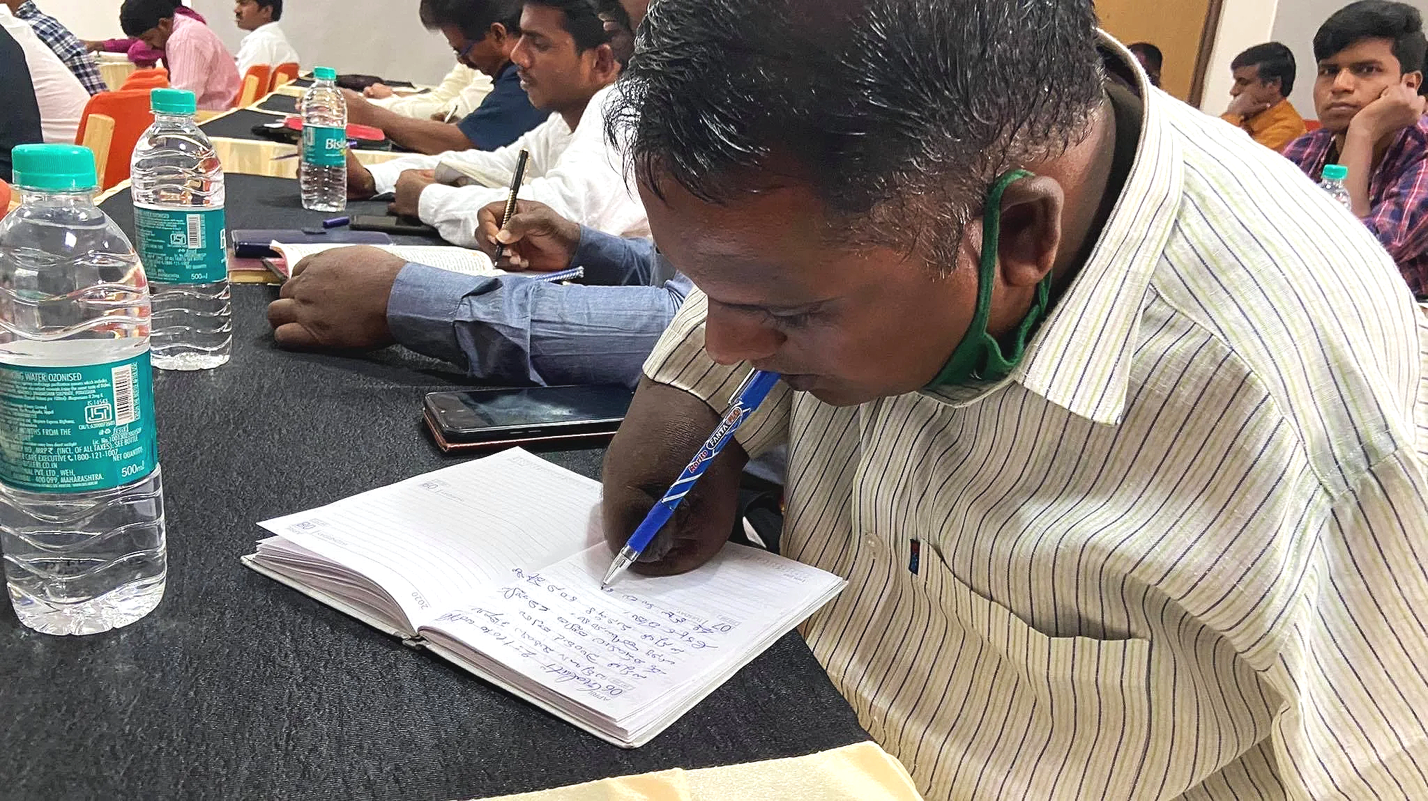 A man in a striped shirt is writing notes in a notebook with a blue pen held in his mouth and supported by an arm, amputated above the elbow during a conference, with other attendees seated around him.