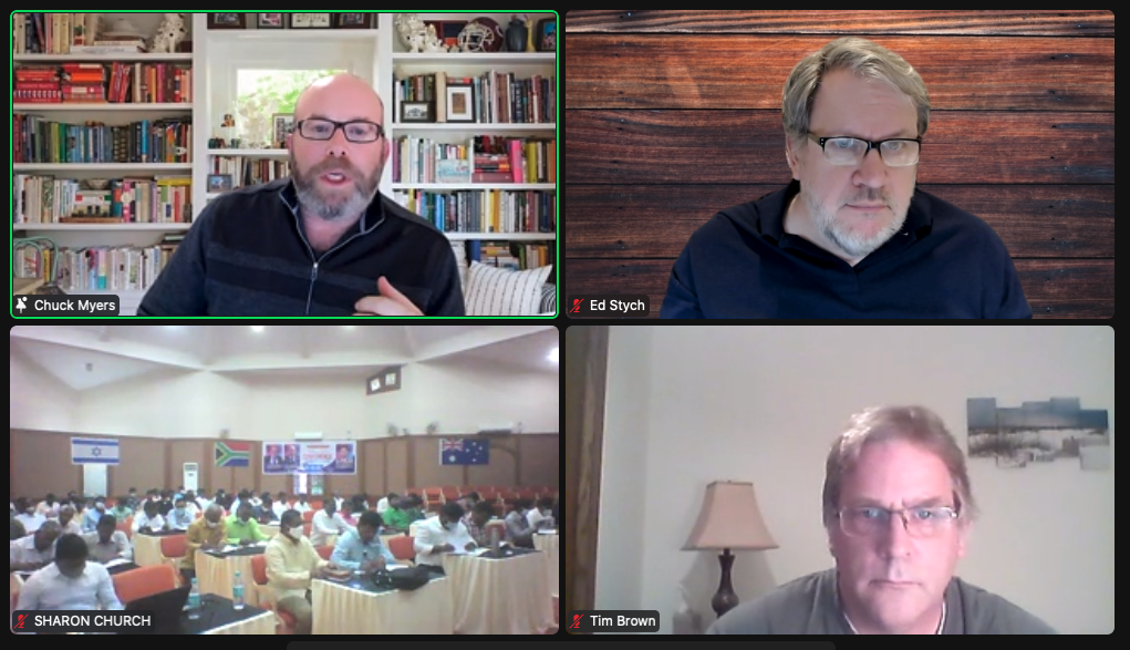 Four individuals participating in a virtual meeting, with a conference room of people in the background, one person speaking, and the others listening or looking at their screens.