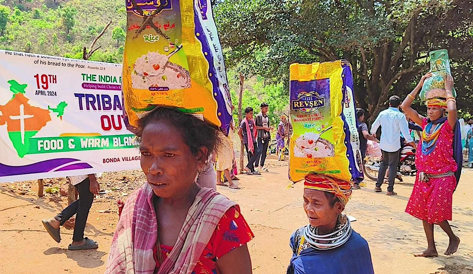 People participating in a community event in a rural area, with large banners advertising food products, and women balancing large bags on their heads, some wearing traditional clothing and jewelry.