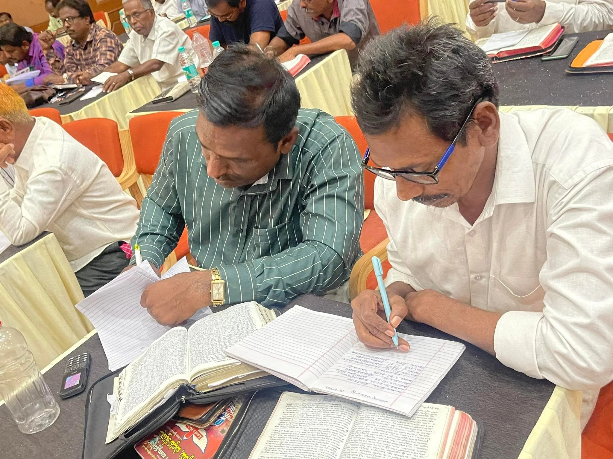 Men sitting at a table in a conference room, reading and taking notes from books and notebooks.
