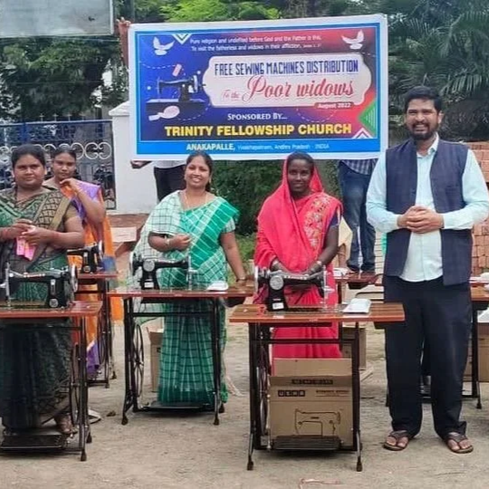 Group of women and a man standing behind sewing machines outdoors, with a large banner behind them advertising free sewing machine distribution for poor widows.