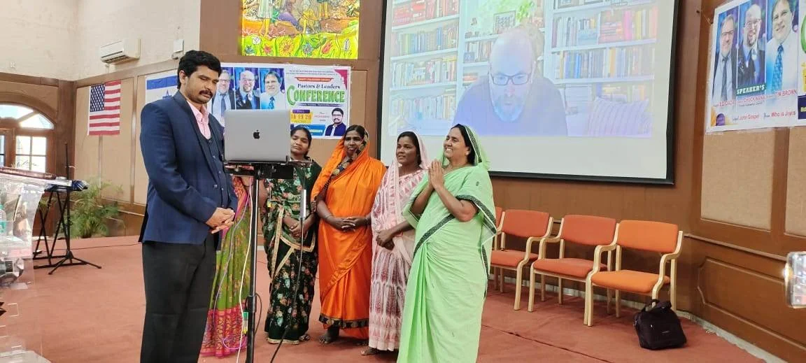Group of women in sarees standing on stage with a man in a suit, participating in a conference or presentation, with a man speaking via video call projected on screen behind them.