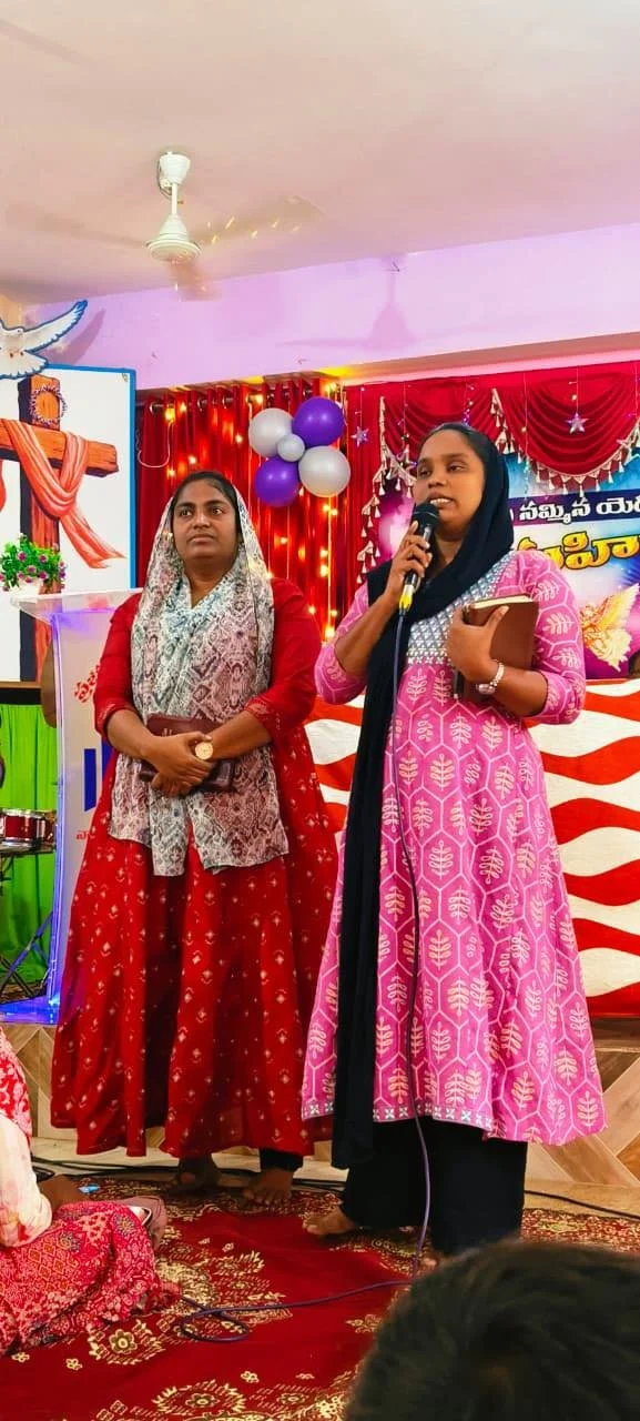 Two women standing on a decorated stage, one holding a microphone and speaking, the other standing beside her. The background features red curtains, balloons, and a banner with Telugu text. The women are wearing traditional Indian dresses.