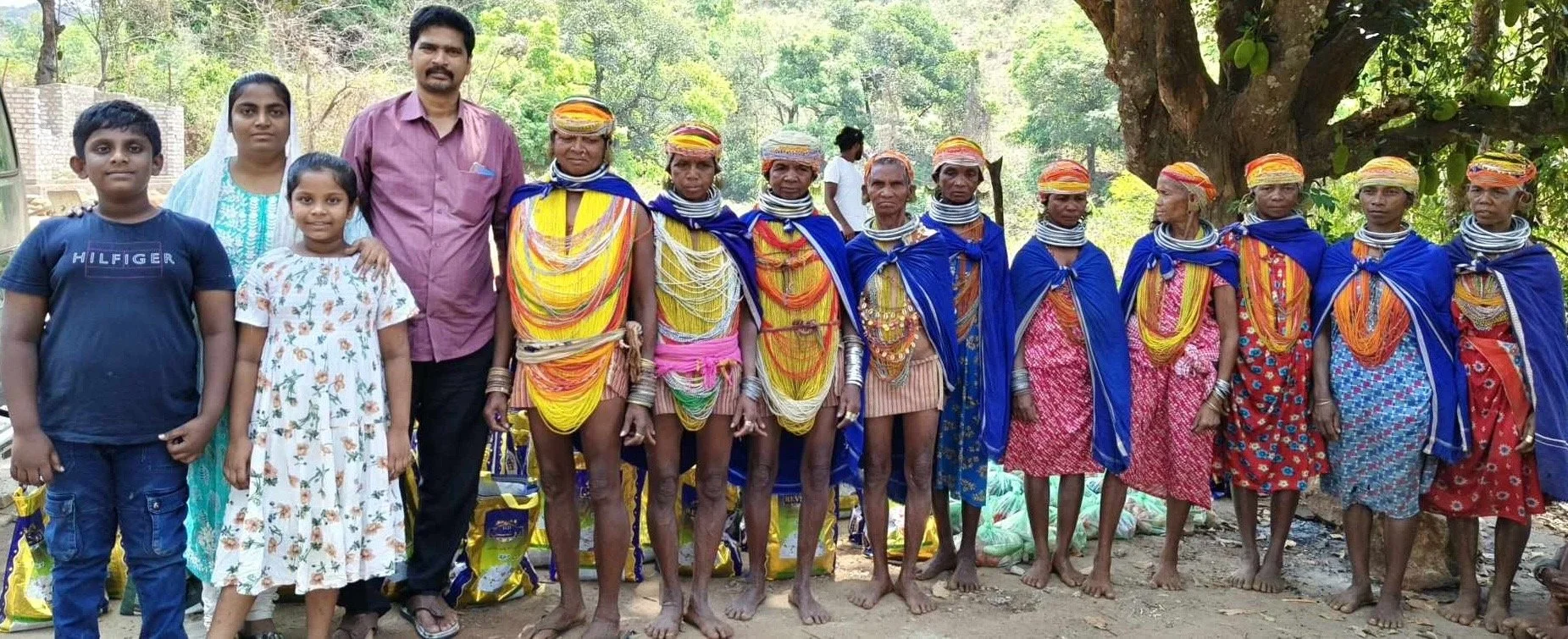 Group of children and elderly women dressed in traditional colorful attire, standing outdoors under a tree with greenery in the background.