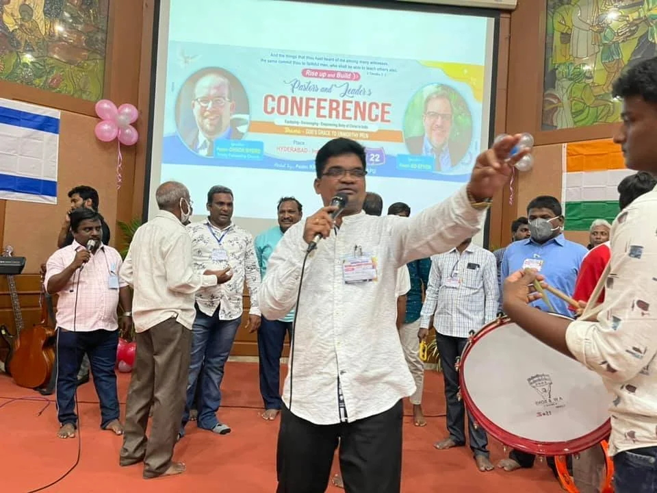 A man speaking into a microphone reaches out to a young person holding a drum during a conference in a hall decorated with balloons and flags, with other people gathered on stage.