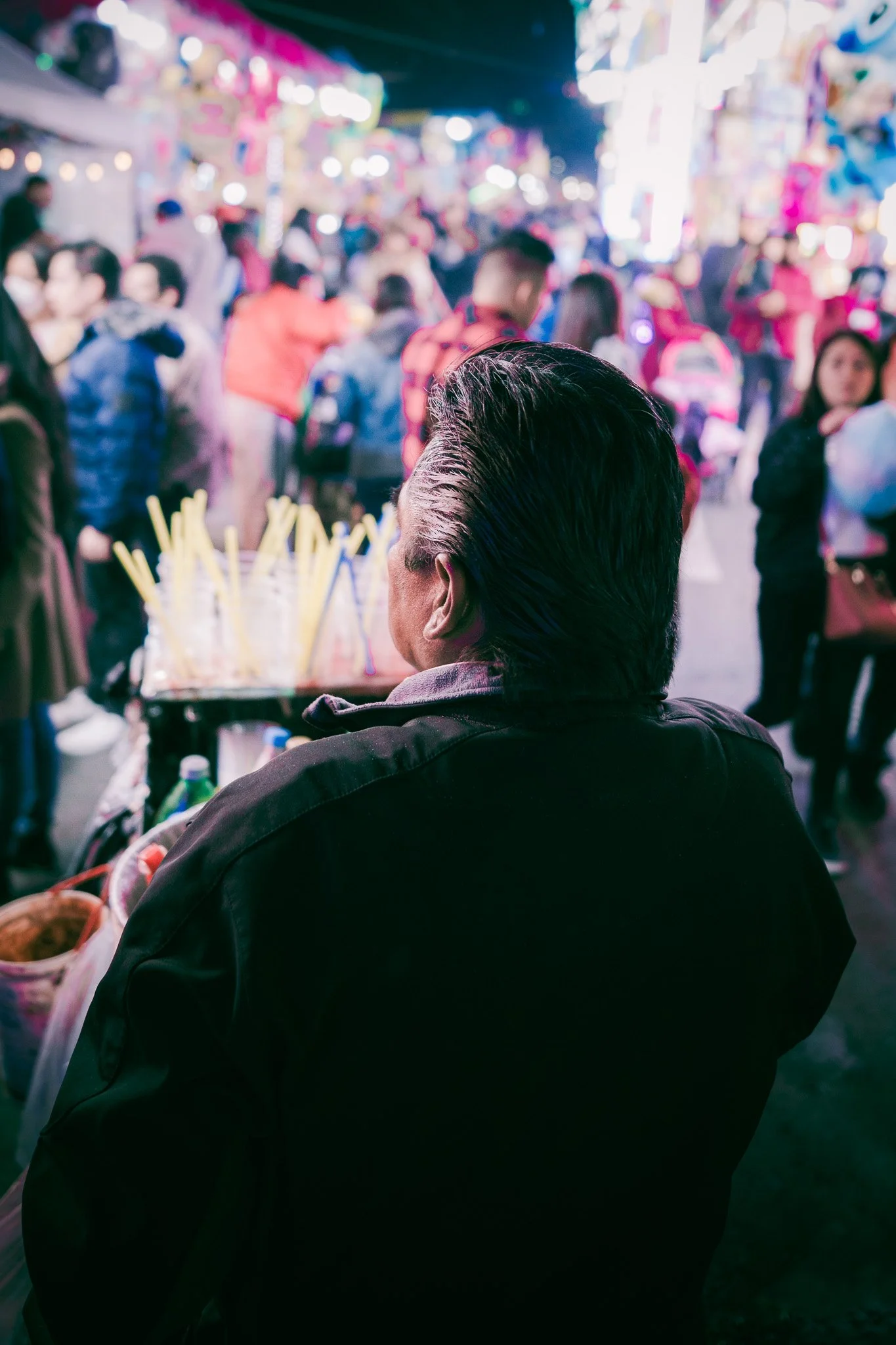 Une personne vue de dos dans un marché animé la nuit, entourée de lumières colorées et de nombreuses personnes