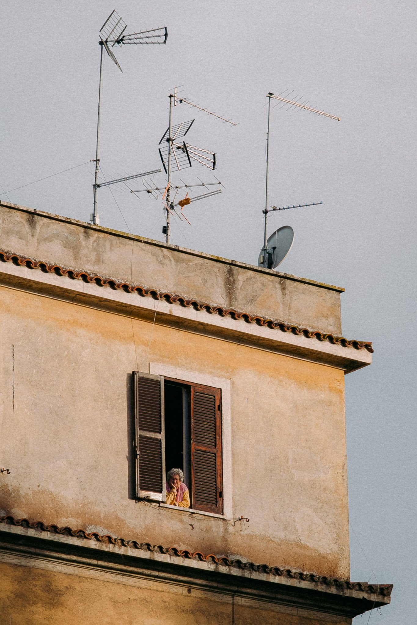 Une vieille femme regarde par une fenêtre ouverte de son appartement au troisième étage. L'immeuble a un toit en tuiles et la façade est beige avec une texture ancienne. Le ciel est gris et plusieurs antennes TV sont installées sur le toit.