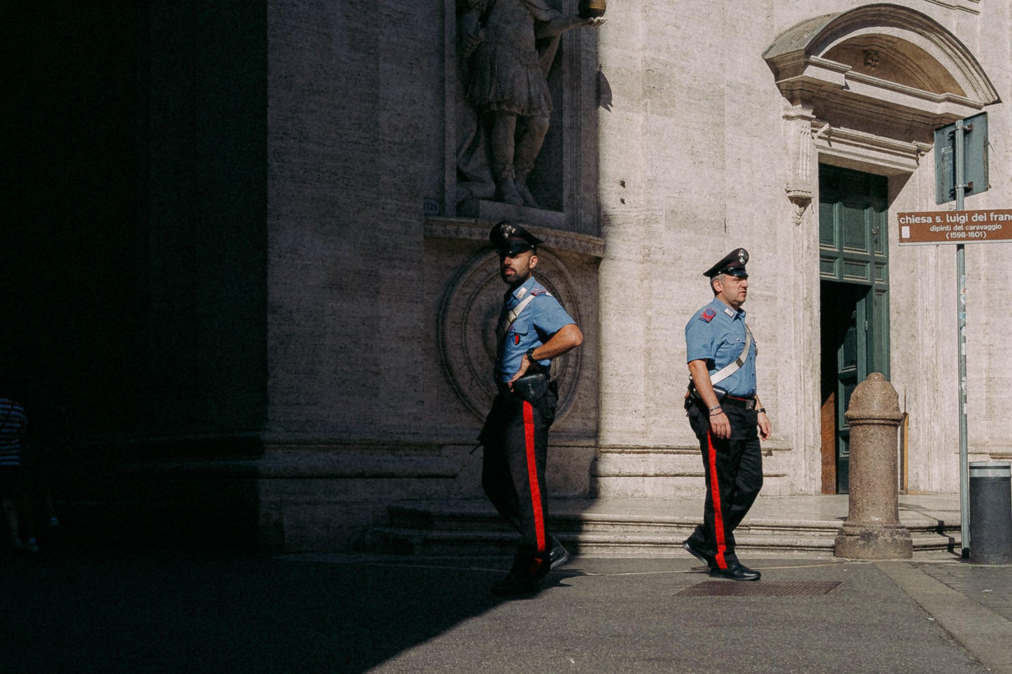 Deux policiers en uniforme déambulent devant une façade en pierre d'une église ou d'un bâtiment historique, avec une sculpture en relief et une plaque d'information, dans un environnement urbain en Italie.