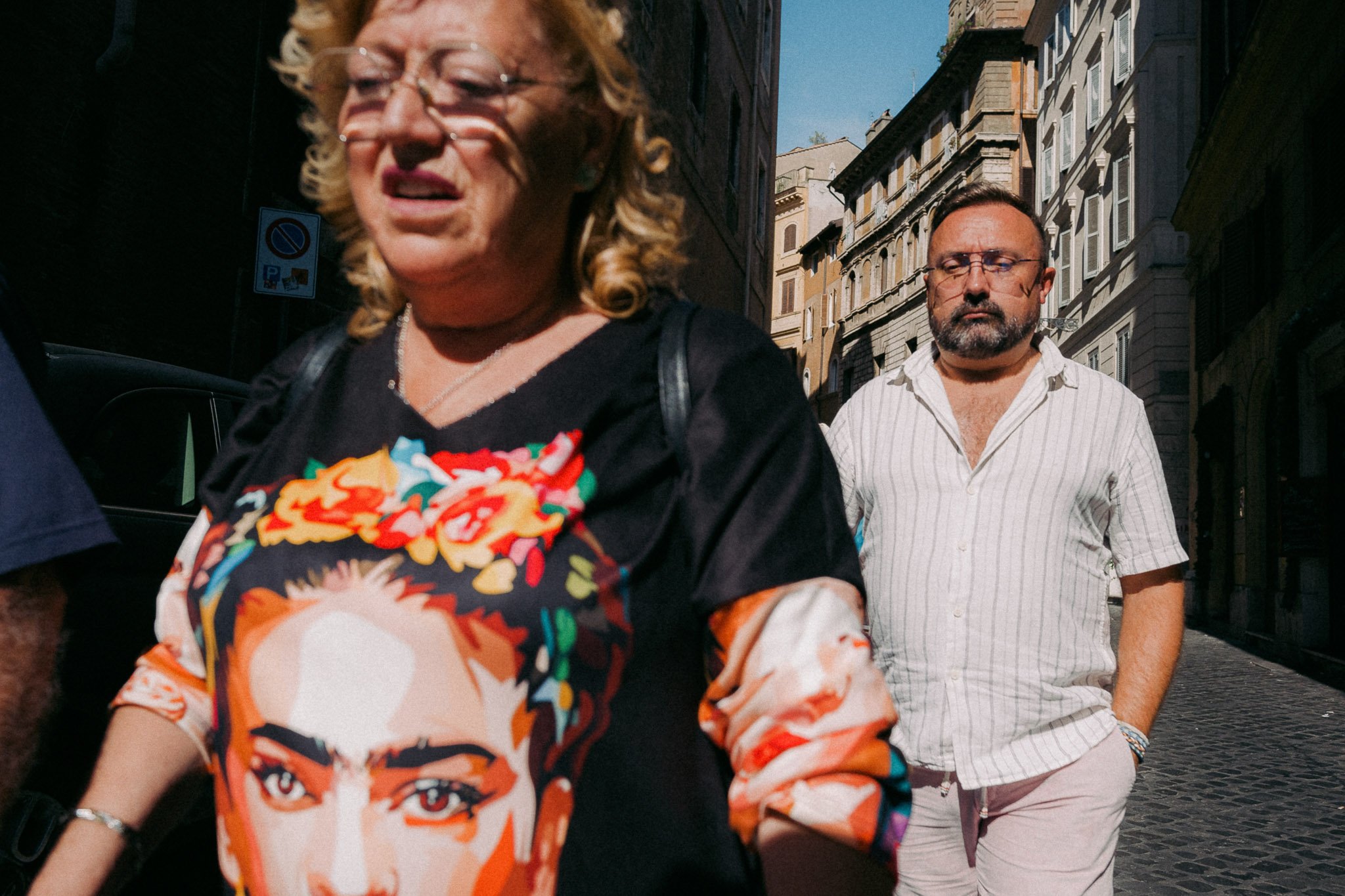 Deux personnes marchent dans une rue urbaine ensoleillée, un homme avec un visage sérieux et une femme avec une chemise colorée avec un visage partiellement visible